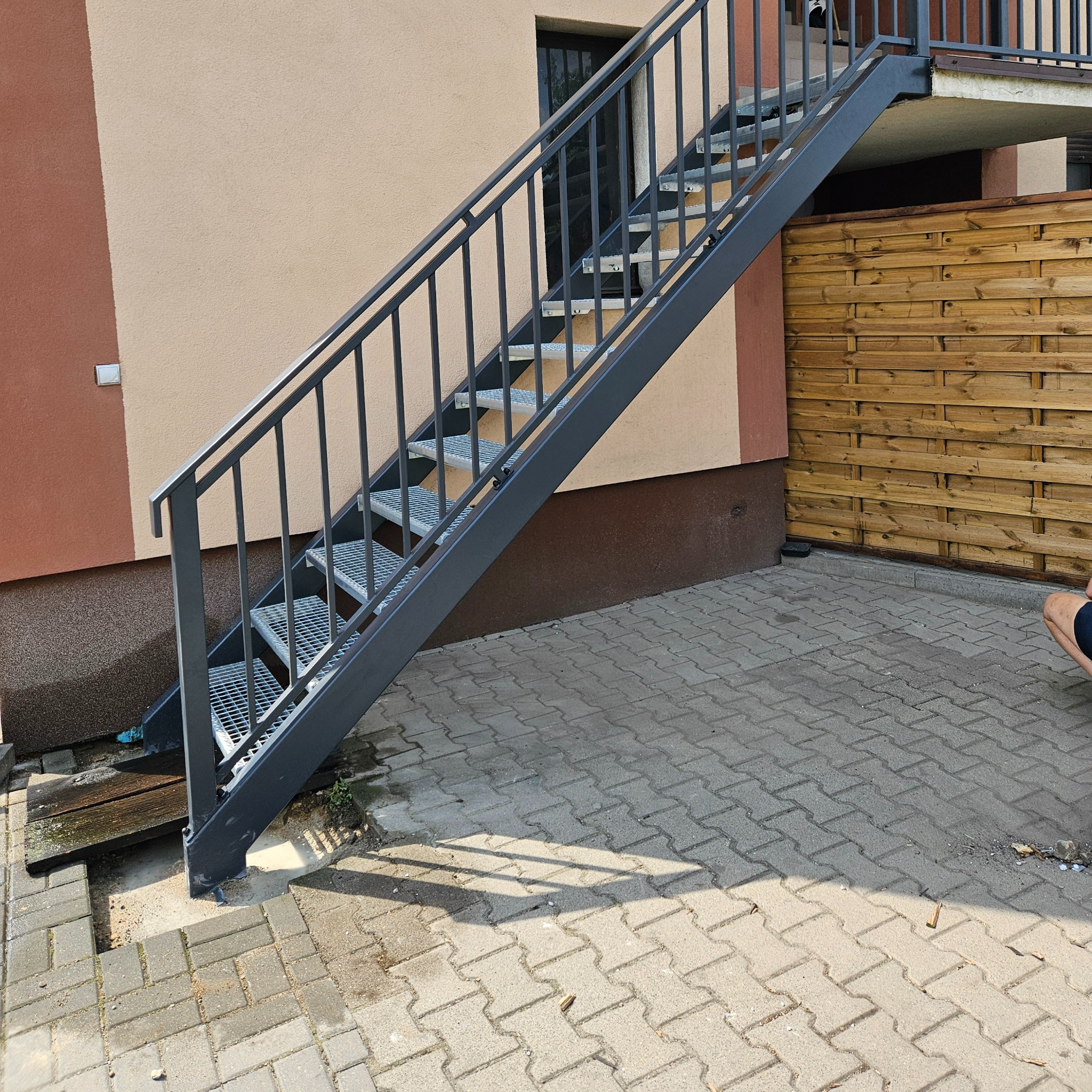 Zewnętrzne schody metalowe z ażurowymi stopniami, prowadzące do drzwi na piętrze, z szarą balustradą i drewnianym płotem w tle. Widok na kostkę brukową.