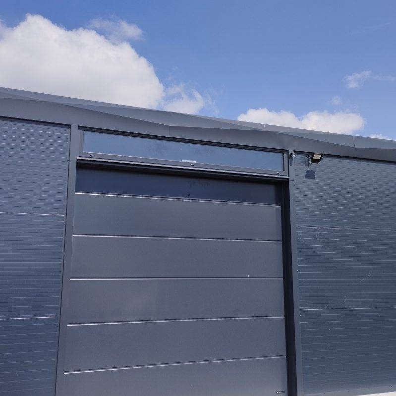 Nowoczesna, segmentowa brama garażowa w odcieniach szarości z poziomymi przetłoczeniami i przeszkleniem u góry, oświetlona punktowym reflektorem, na tle błękitnego nieba.