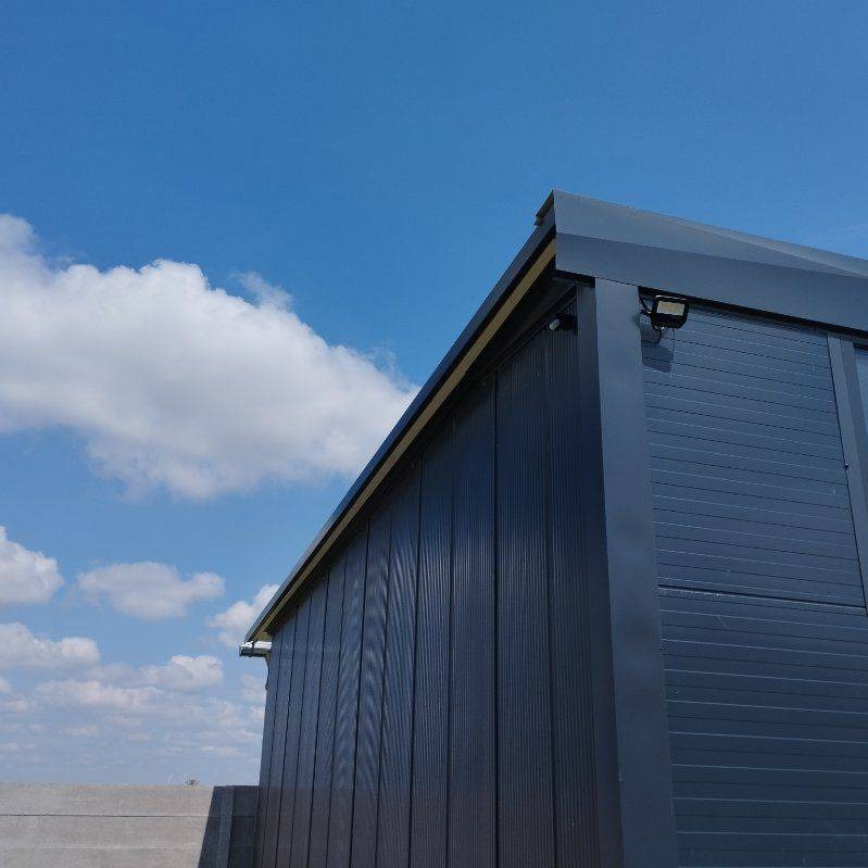 Nowoczesna, grafitowa zabudowa balkonu z metalu, widziana z dołu, z widocznym oświetleniem i fragmentem betonowego murku na tle błękitnego nieba z chmurami.