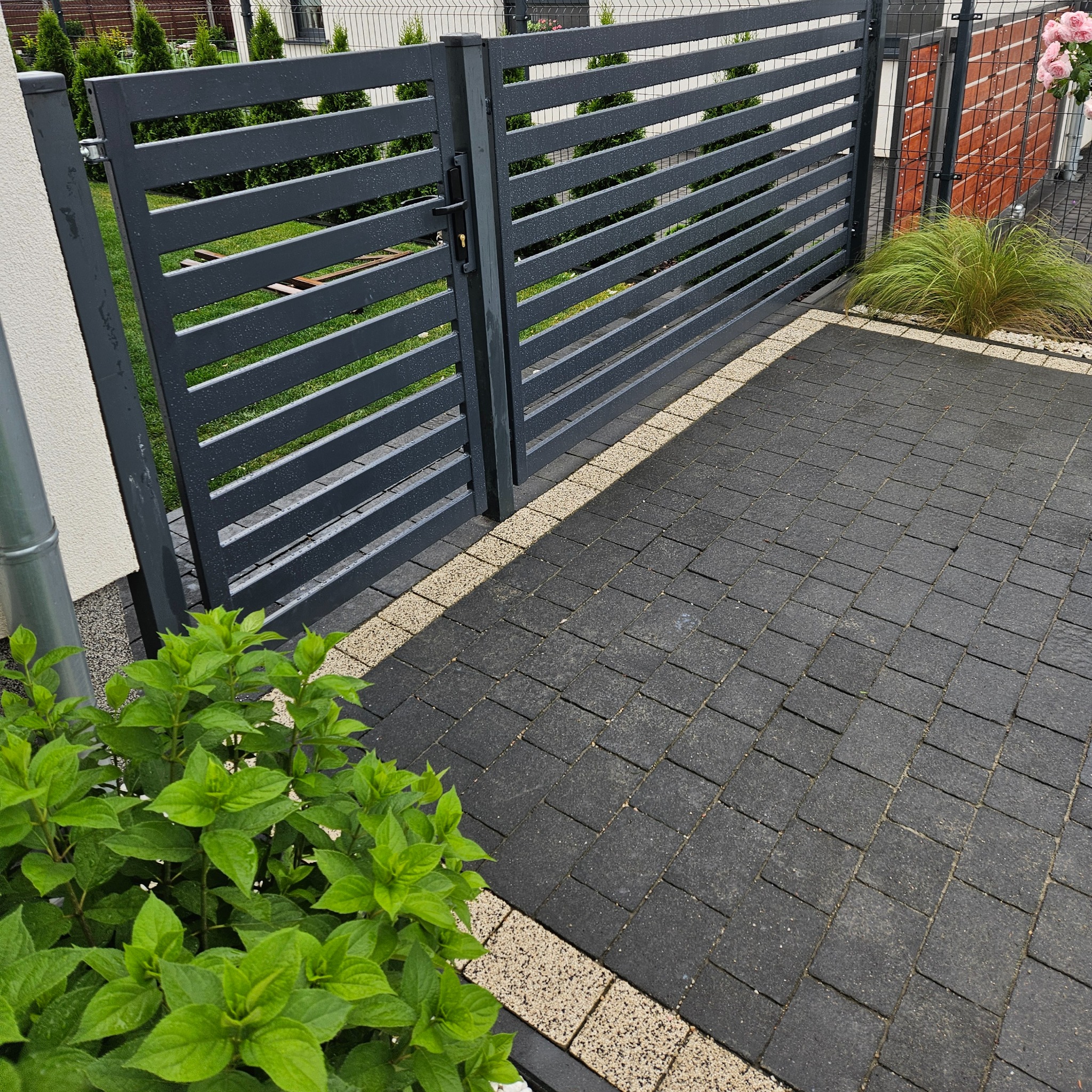 Nowoczesna brama wjazdowa z poziomych profili, antracytowa, na tle kostki brukowej i zieleni. Minimalistyczny design, detal zamykania.