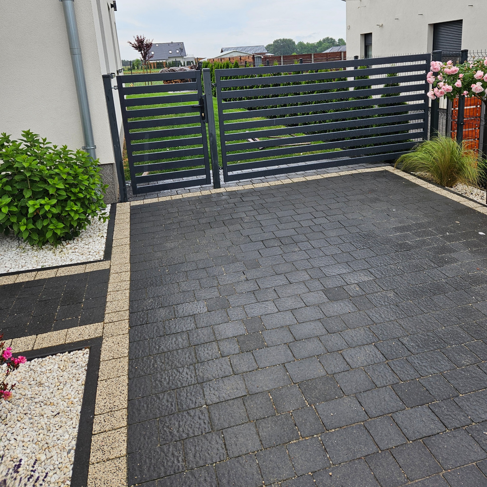 Nowoczesna, pozioma brama wjazdowa z szarego metalu, z widoczną kostką brukową i elementami dekoracyjnymi z kamienia i roślin. Minimalistyczny design.
