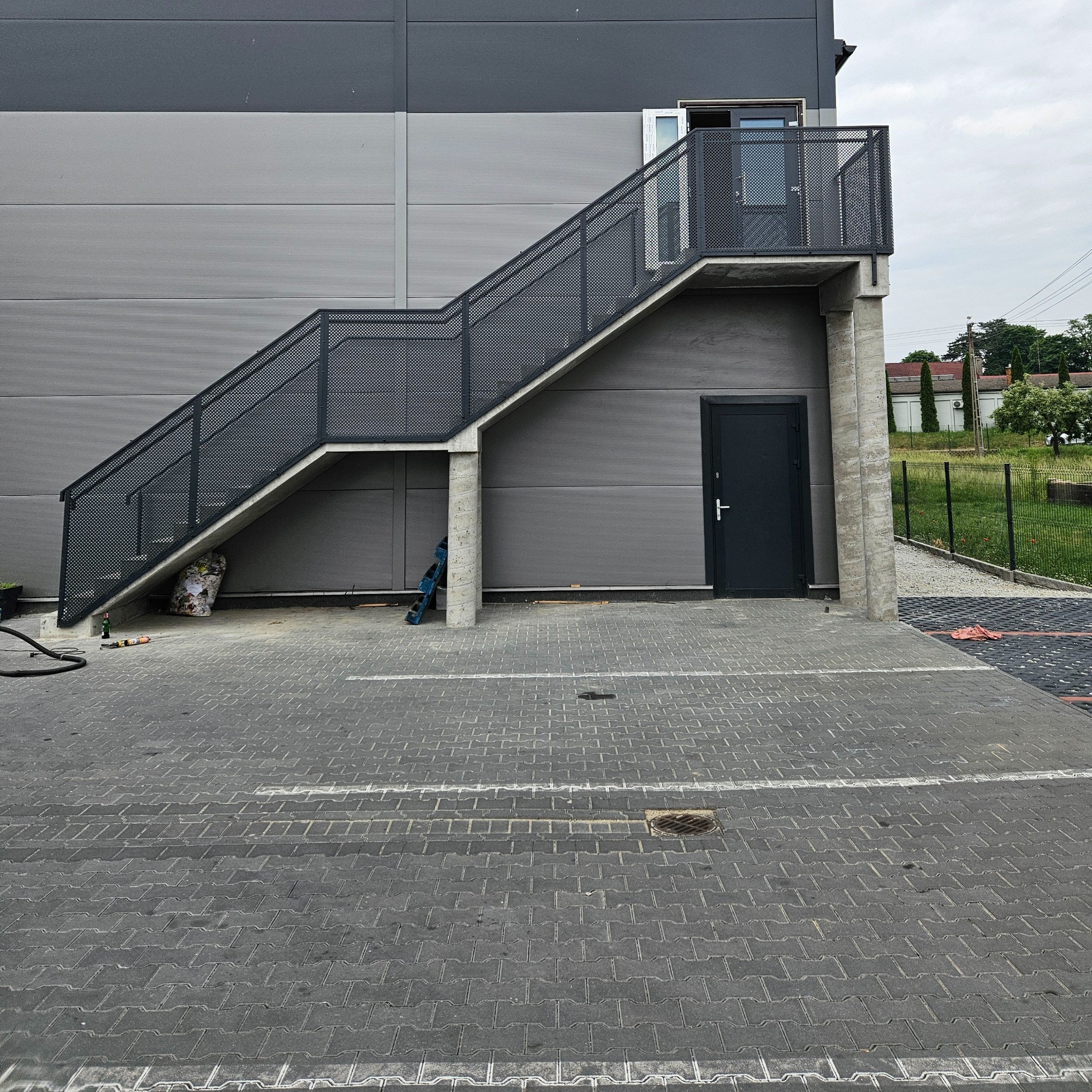 Zewnętrzne schody metalowe z perforowaną balustradą prowadzą do drzwi na piętrze szarego budynku przemysłowego. Widoczne narzędzia i materiały budowlane.