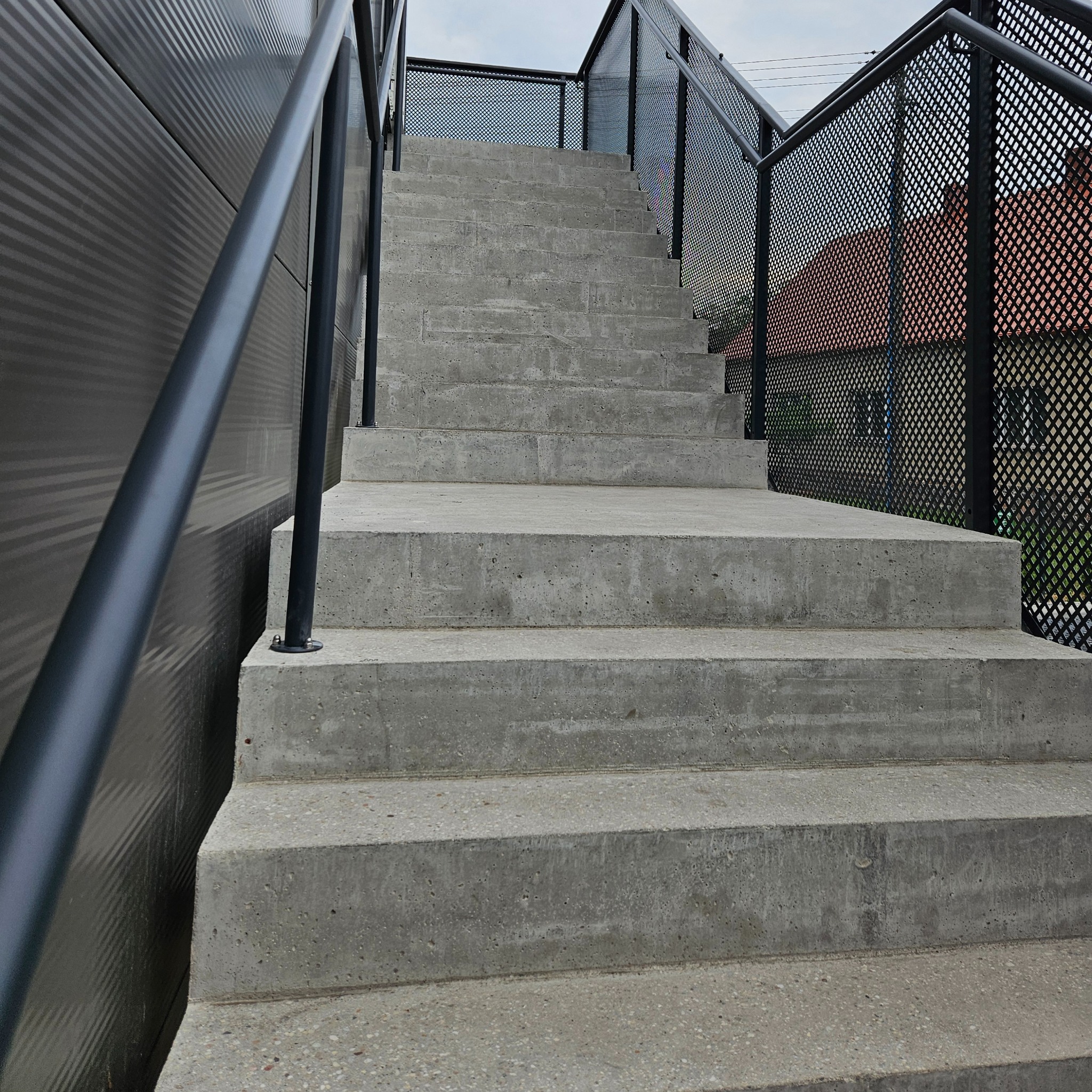 Betonowe schody zewnętrzne z czarną, metalową balustradą o ażurowym wzorze, prowadzące w górę. Widok z dołu, z boku budynku z blachy trapezowej.