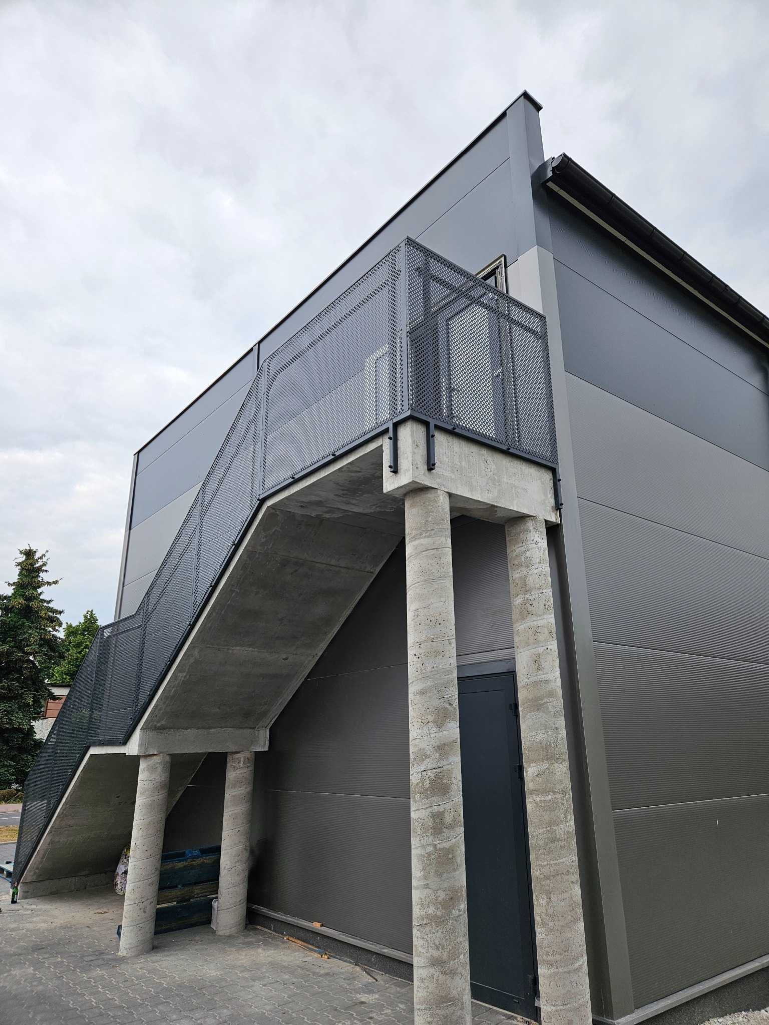 Zewnętrzne schody betonowe z metalową ażurową balustradą prowadzące do budynku o szarej elewacji. Widok z dołu, pochmurne niebo w tle.