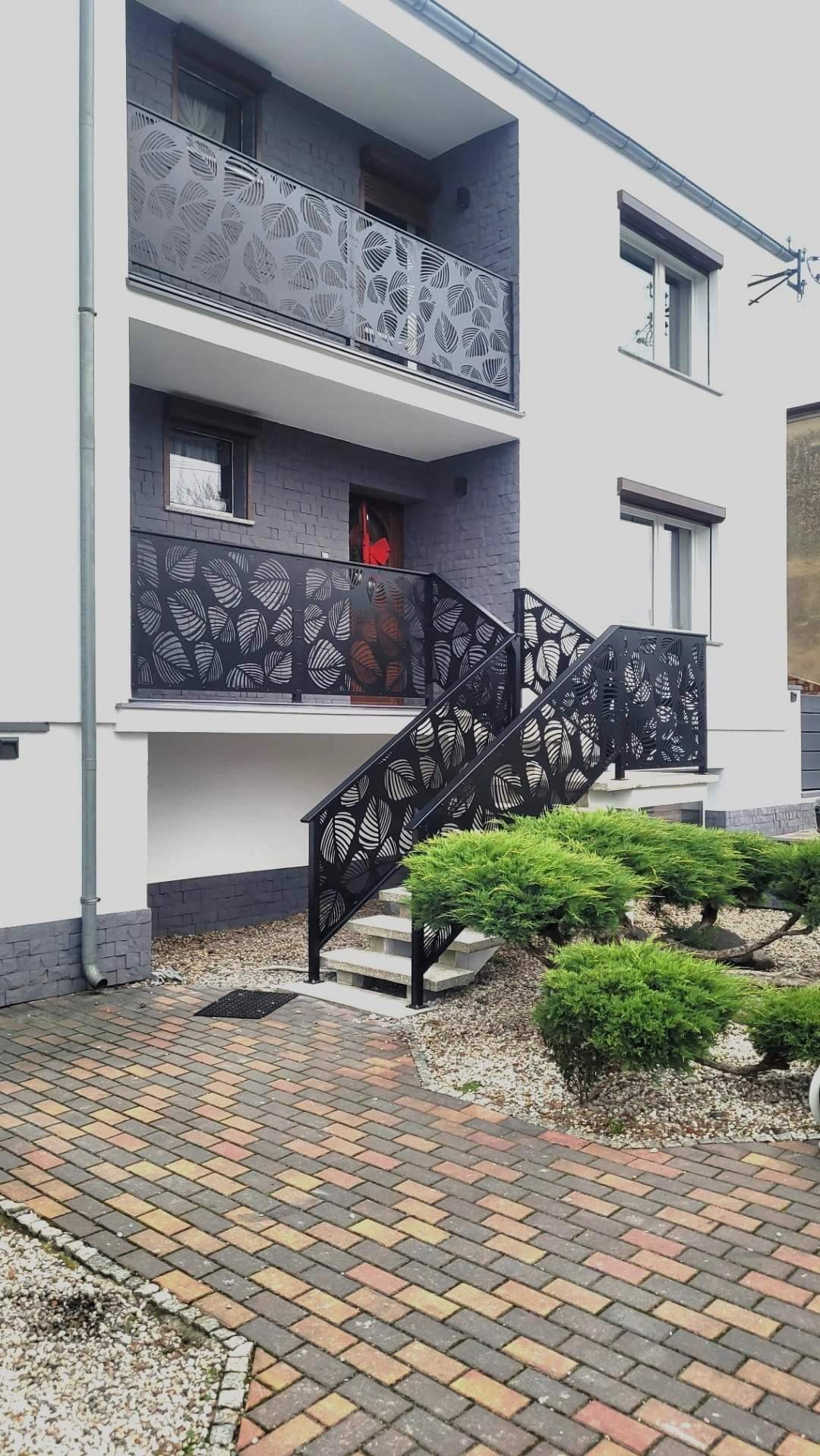 Nowoczesna balustrada balkonowa i schodowa z ażurowym wzorem liści, czarna, na tle białej elewacji z szarym akcentem, widok z poziomu chodnika.