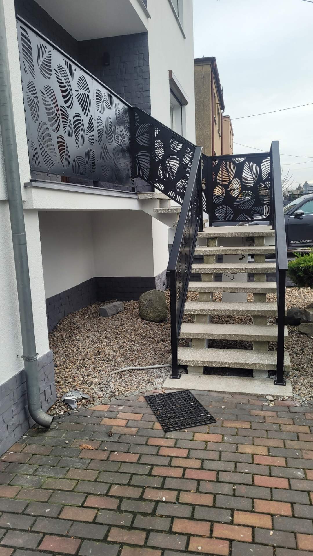 Betonowe schody zewnętrzne z czarną, ażurową balustradą o wzorze liści, prowadzące na podwyższony taras. Ciekawy detal: kamień dekoracyjny obok schodów.