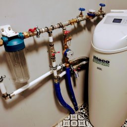 INNOVA Jakub Paszkowski - System uzdatniania wody Viteco Smart Rain 20L z filtrem wstępnym, zaworami i rurami zamontowany na ścianie nad ozdobną podłogą.