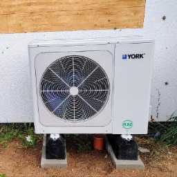 Biała pompa ciepła marki York, model z czynnikiem chłodniczym R32, umieszczona na betonowych podstawach przy ścianie budynku z fakturą baranka i drewnianym wykończeniem.