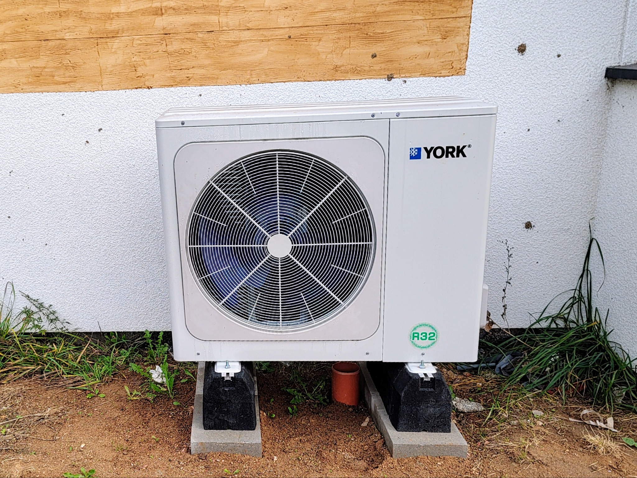 Biała pompa ciepła marki York, model z czynnikiem chłodniczym R32, umieszczona na betonowych podstawach przy ścianie budynku z fakturą baranka i drewnianym wykończeniem.