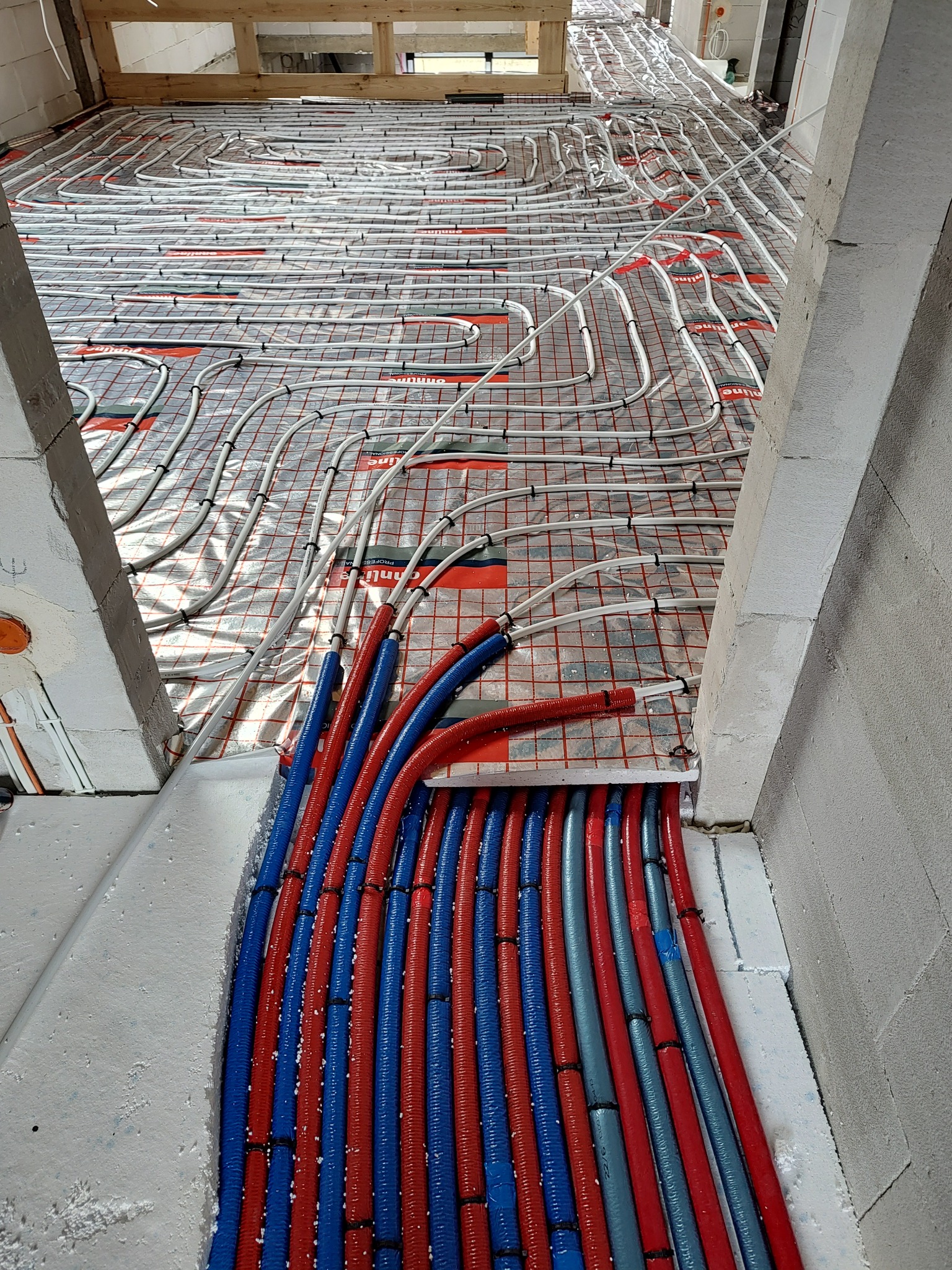 Rozłożone rury ogrzewania podłogowego na siatce z folią odblaskową, widoczne niebieskie i czerwone rury doprowadzające, surowy stan budowy.