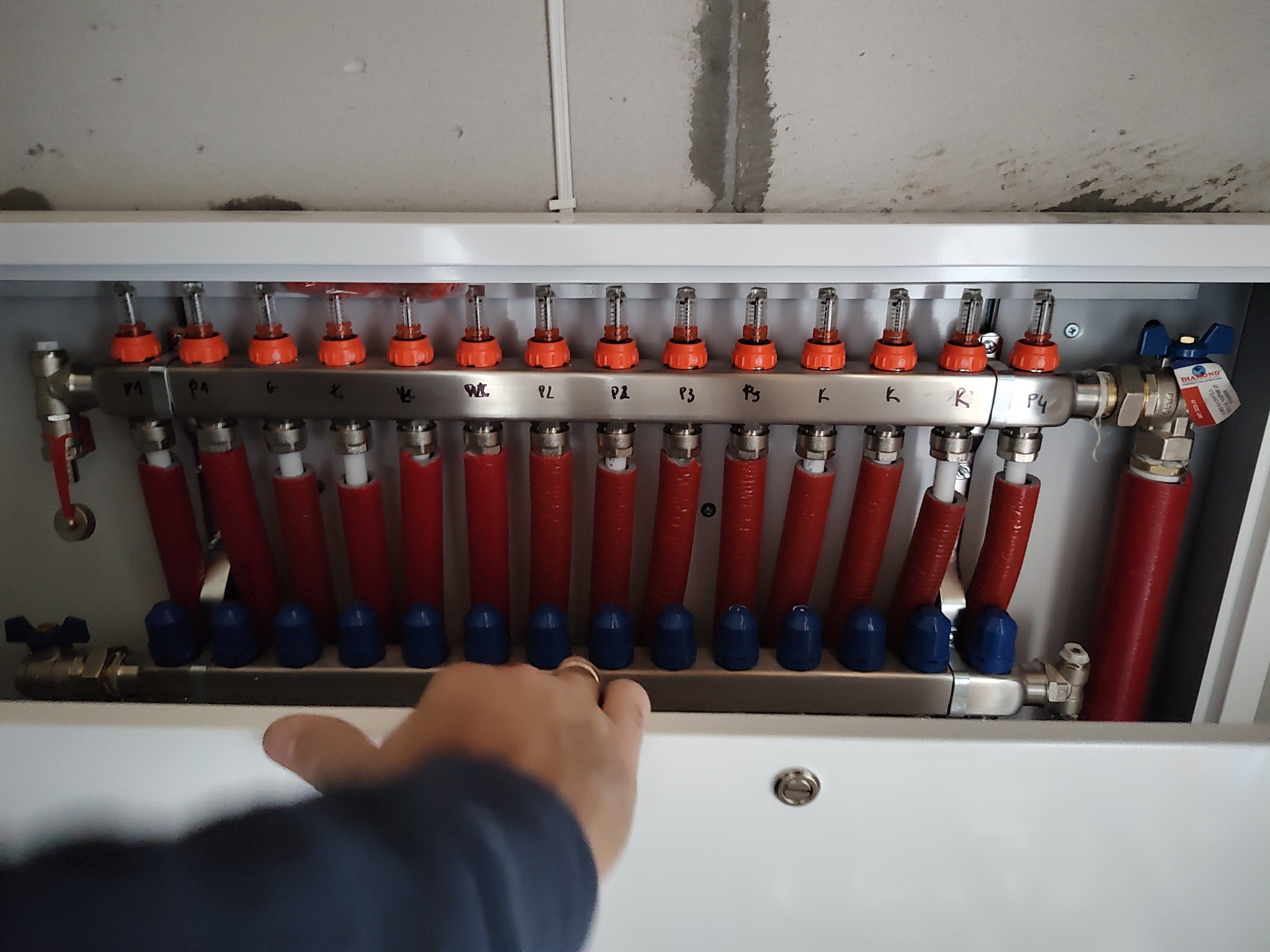 Rozdzielacz ogrzewania podłogowego w szafce, z czerwonymi rurami i niebieskimi zaworami, oznaczony symbolami WL, PL, P1, P2, P3, K, P4, regulowany dłonią w złotym pierścionku.