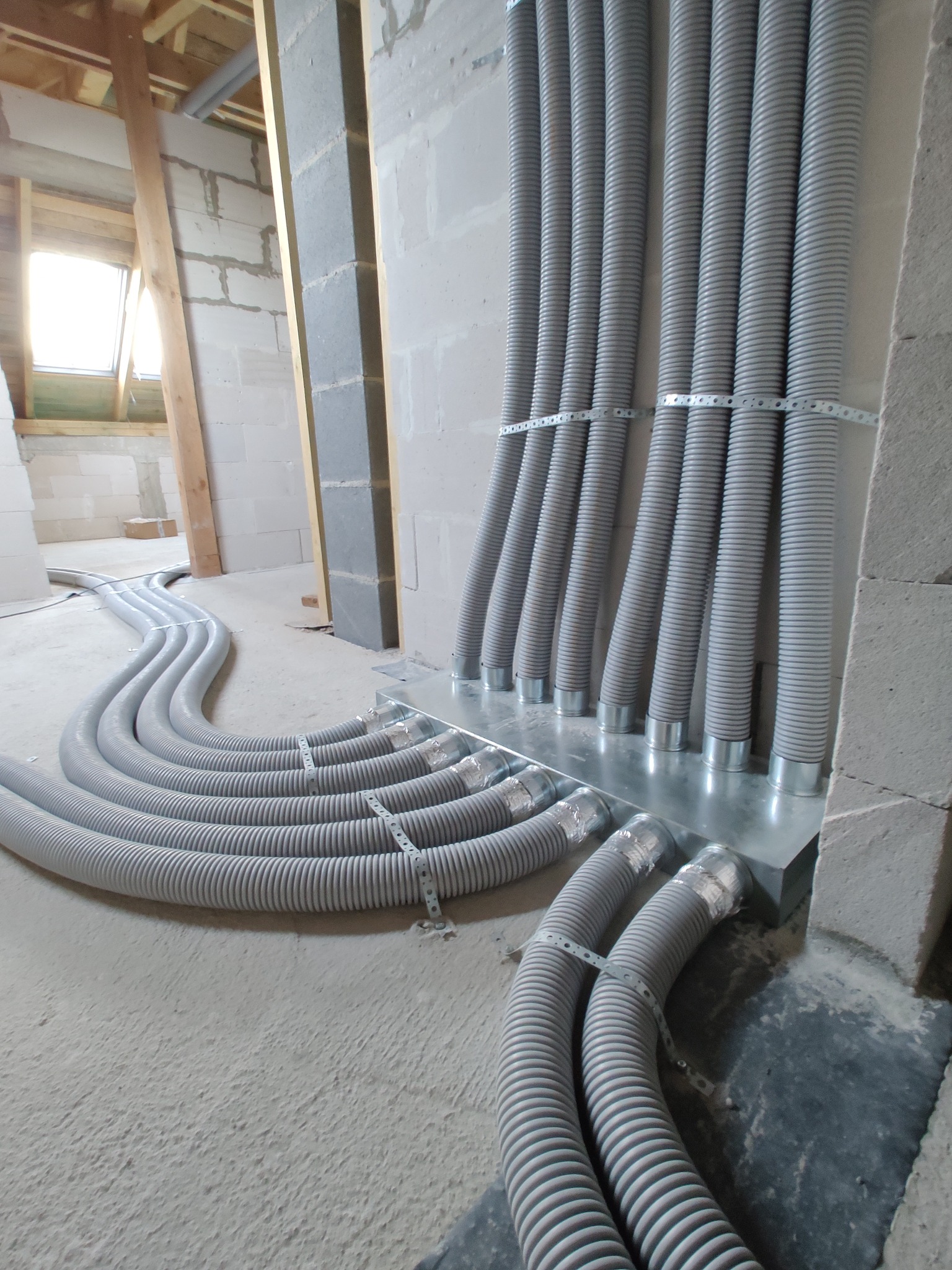 Szare, karbowane rury wentylacyjne ułożone na podłodze i przymocowane do ściany na metalowej płycie, widoczne w surowym wnętrzu budynku w trakcie budowy.
