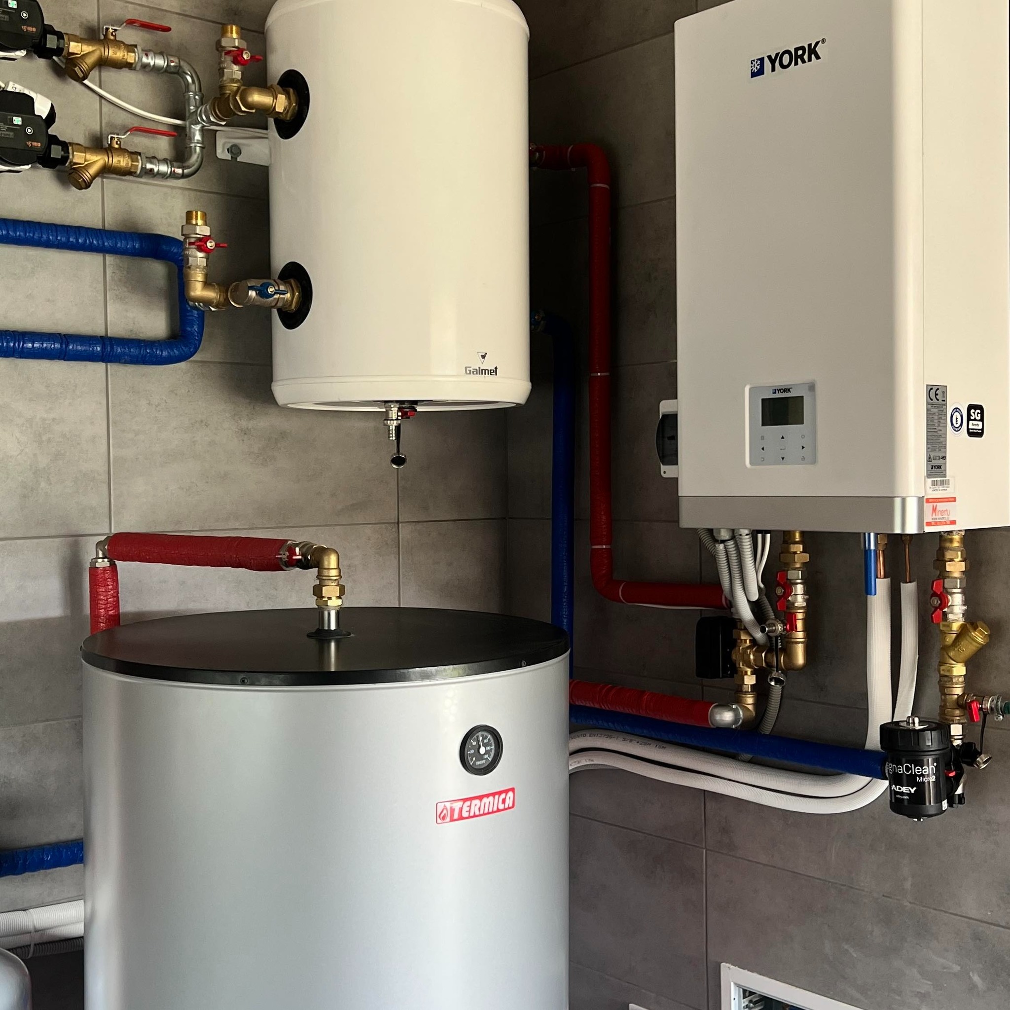 Dwa białe zasobniki ciepłej wody użytkowej marki Galmet i Termica, połączone rurami w izolacji czerwonej i niebieskiej, obok wiszący piec grzewczy marki York, na tle szarej ściany z płytek.