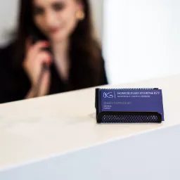 Wizytówki firmy prawniczej w metalowym podajniku na białym biurku, w tle rozmyta postać kobiety rozmawiającej przez telefon.