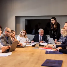 Zespół prawników podczas spotkania w sali konferencyjnej, z laptopami i notatnikami na drewnianym stole, kobieta w czarnej sukience stoi i prezentuje coś grupie.