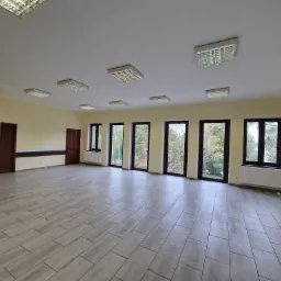 Duże, puste pomieszczenie z szarą podłogą imitującą drewno, jasnymi ścianami, ciemnymi oknami i sufitowymi lampami rastrowymi.