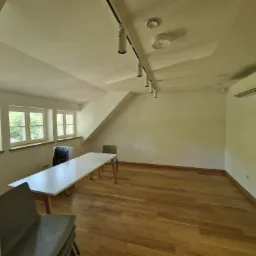 Pusta sala z drewnianą podłogą, białymi ścianami, oknami z widokiem na zieleń, oświetleniem szynowym na suficie i klimatyzacją na ścianie, przygotowana do użytku.