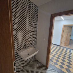 Spread Joy - Umywalka zamontowana na ścianie wyłożonej czarno-białą mozaiką, widok z perspektywy wejścia do łazienki, podłoga również wyłożona mozaiką, w tle widoczna podłoga w szachownicę.