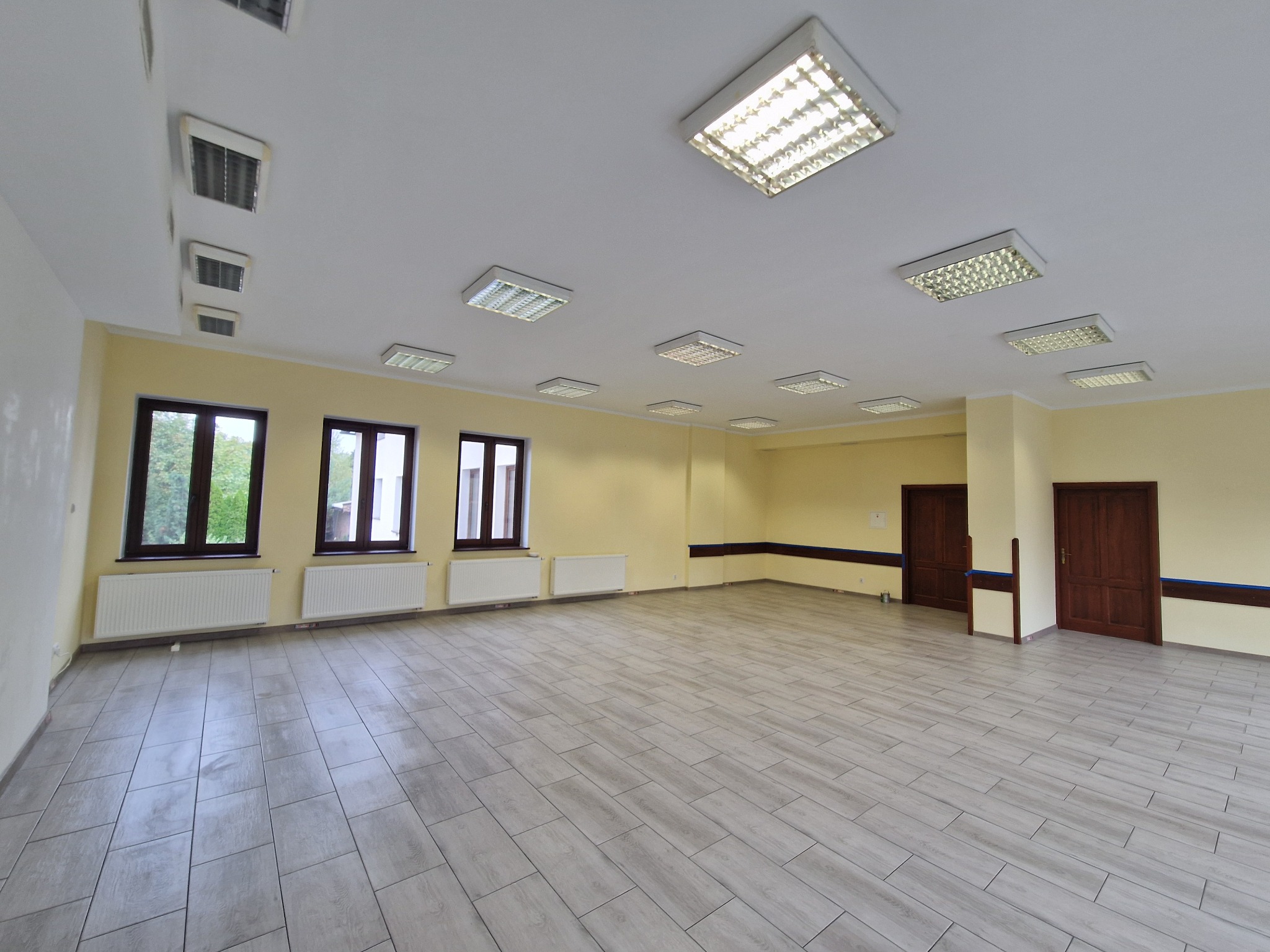 Pusta, jasna sala z nową, szarą podłogą imitującą panele, żółte ściany, białe grzejniki pod trzema oknami i liczne kwadratowe lampy sufitowe.