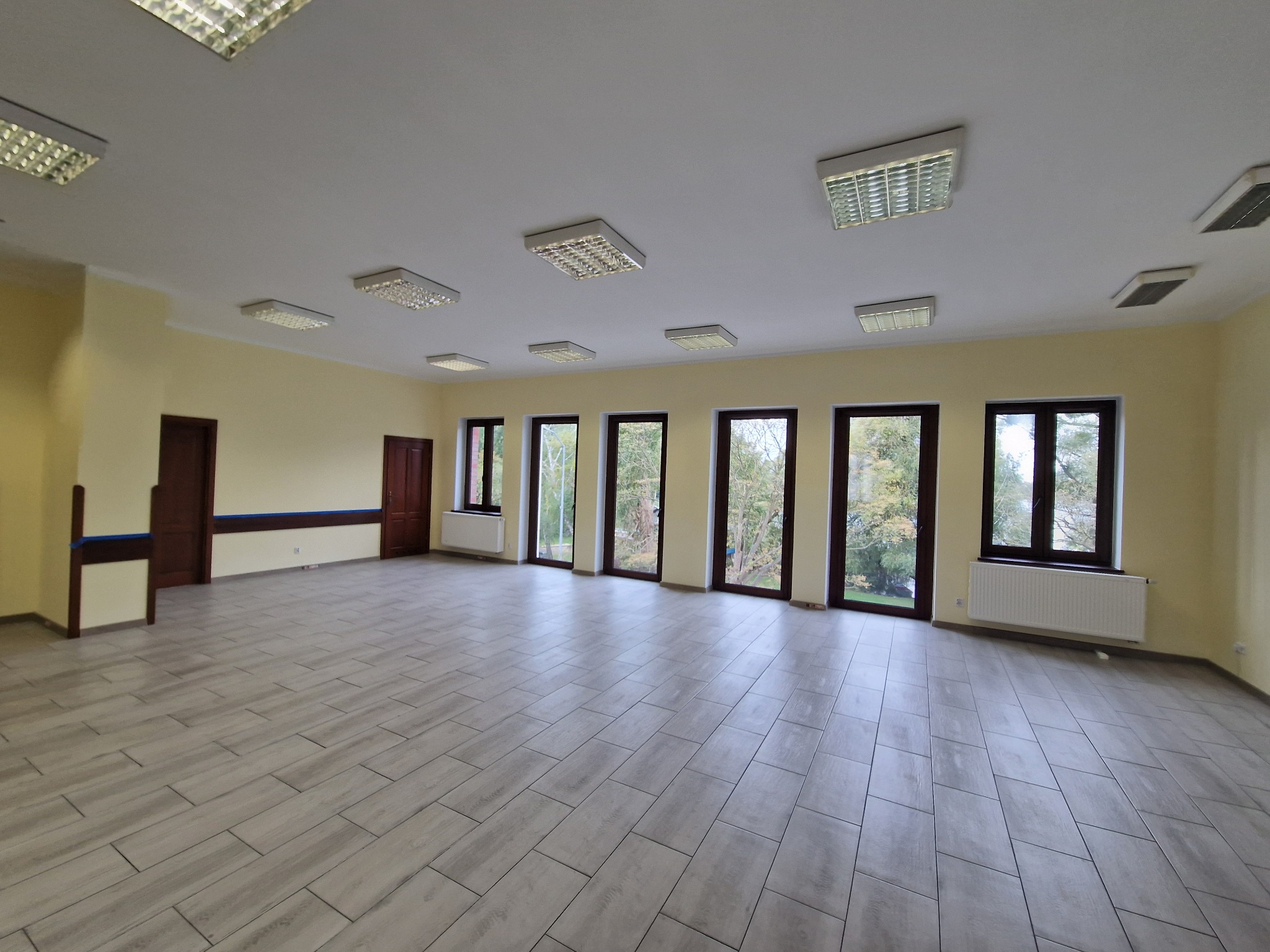 Duże, puste pomieszczenie z szarą podłogą imitującą drewno, jasnymi ścianami, ciemnymi oknami i sufitowymi lampami rastrowymi.