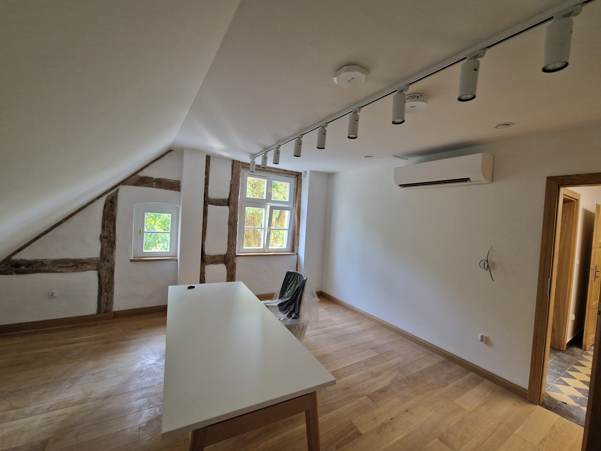 Pokój po remoncie: drewniana podłoga, białe ściany z fragmentami muru pruskiego, nowoczesne oświetlenie szynowe i klimatyzacja, minimalistyczny stół i krzesło zabezpieczone folią.