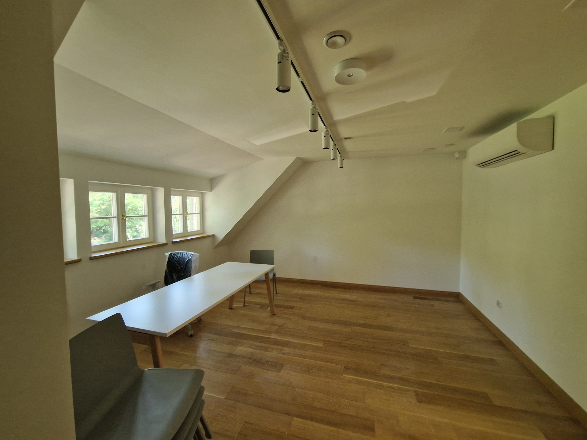 Pusta sala z drewnianą podłogą, białymi ścianami, oknami z widokiem na zieleń, oświetleniem szynowym na suficie i klimatyzacją na ścianie, przygotowana do użytku.