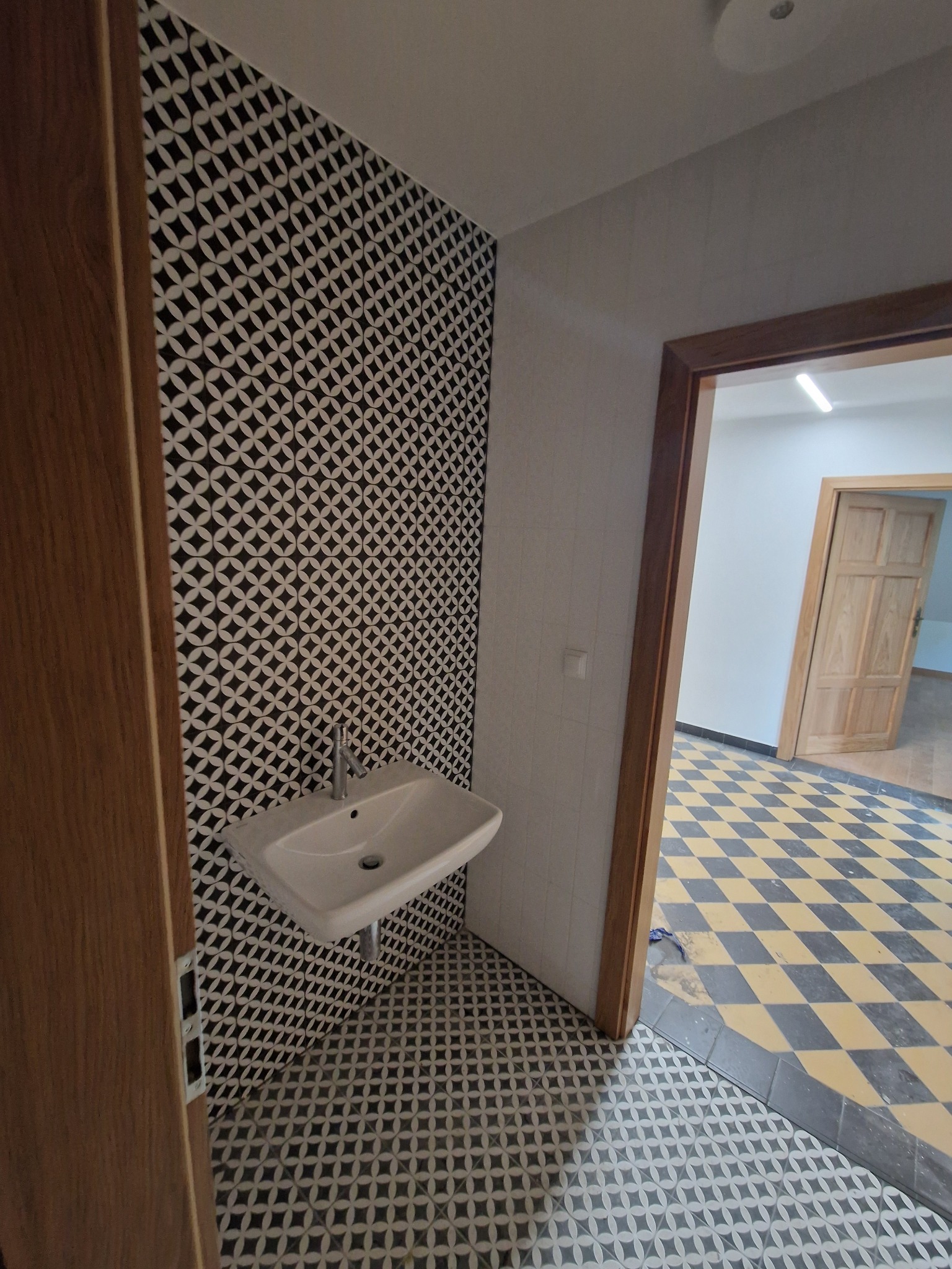 Umywalka zamontowana na ścianie wyłożonej czarno-białą mozaiką, widok z perspektywy wejścia do łazienki, podłoga również wyłożona mozaiką, w tle widoczna podłoga w szachownicę.
