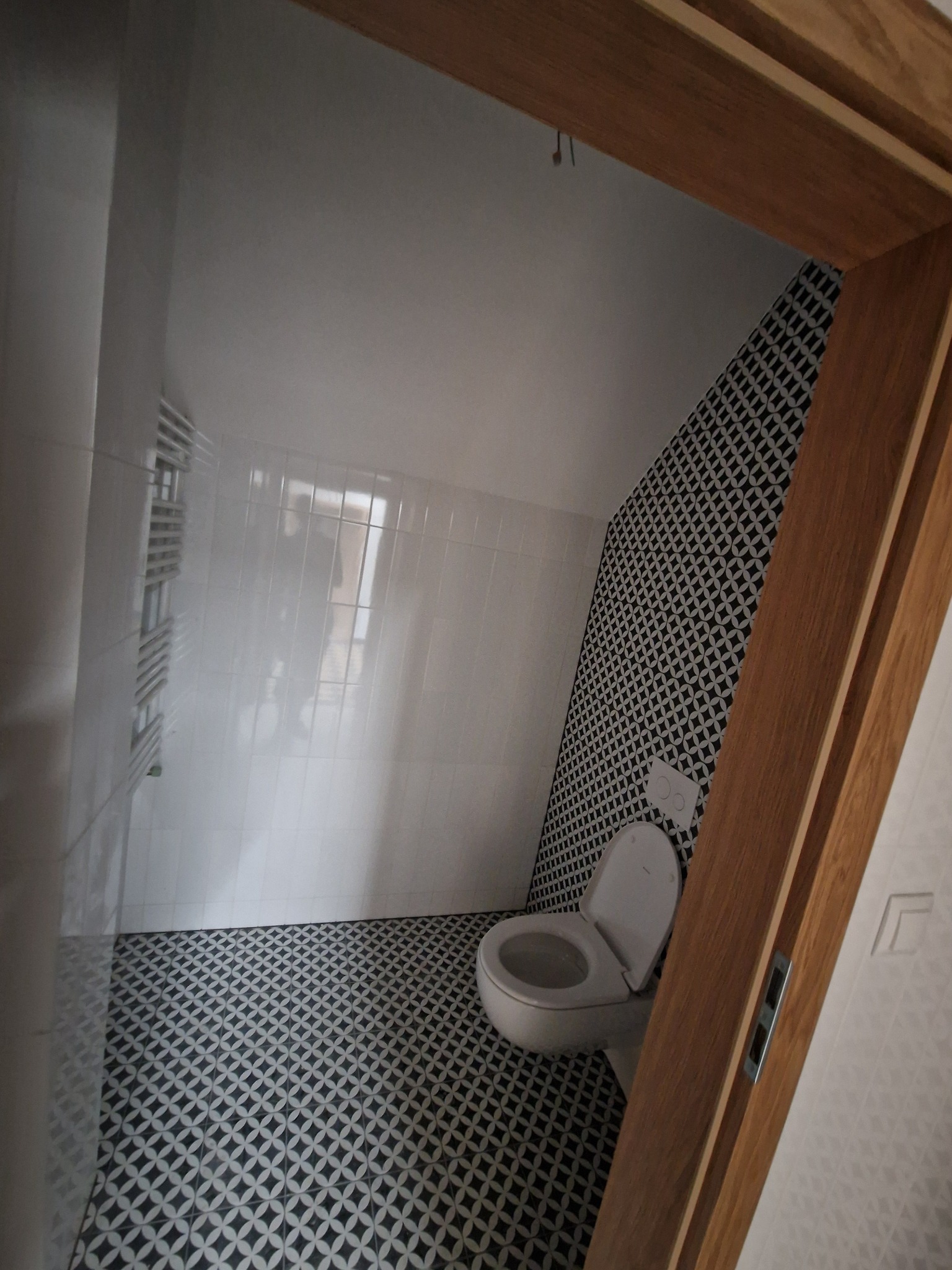 Wykończona łazienka z czarno-białą mozaiką na podłodze i ścianie, białe płytki na pozostałych ścianach, toaleta wisząca oraz grzejnik drabinkowy, widok przez drewnianą futrynę.