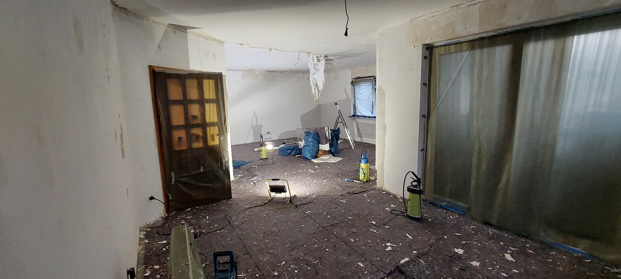 Pokój w trakcie prac przygotowawczych do remontu: zabezpieczone folią drzwi, oświetlenie robocze, worki na gruz i resztki tapet na podłodze.