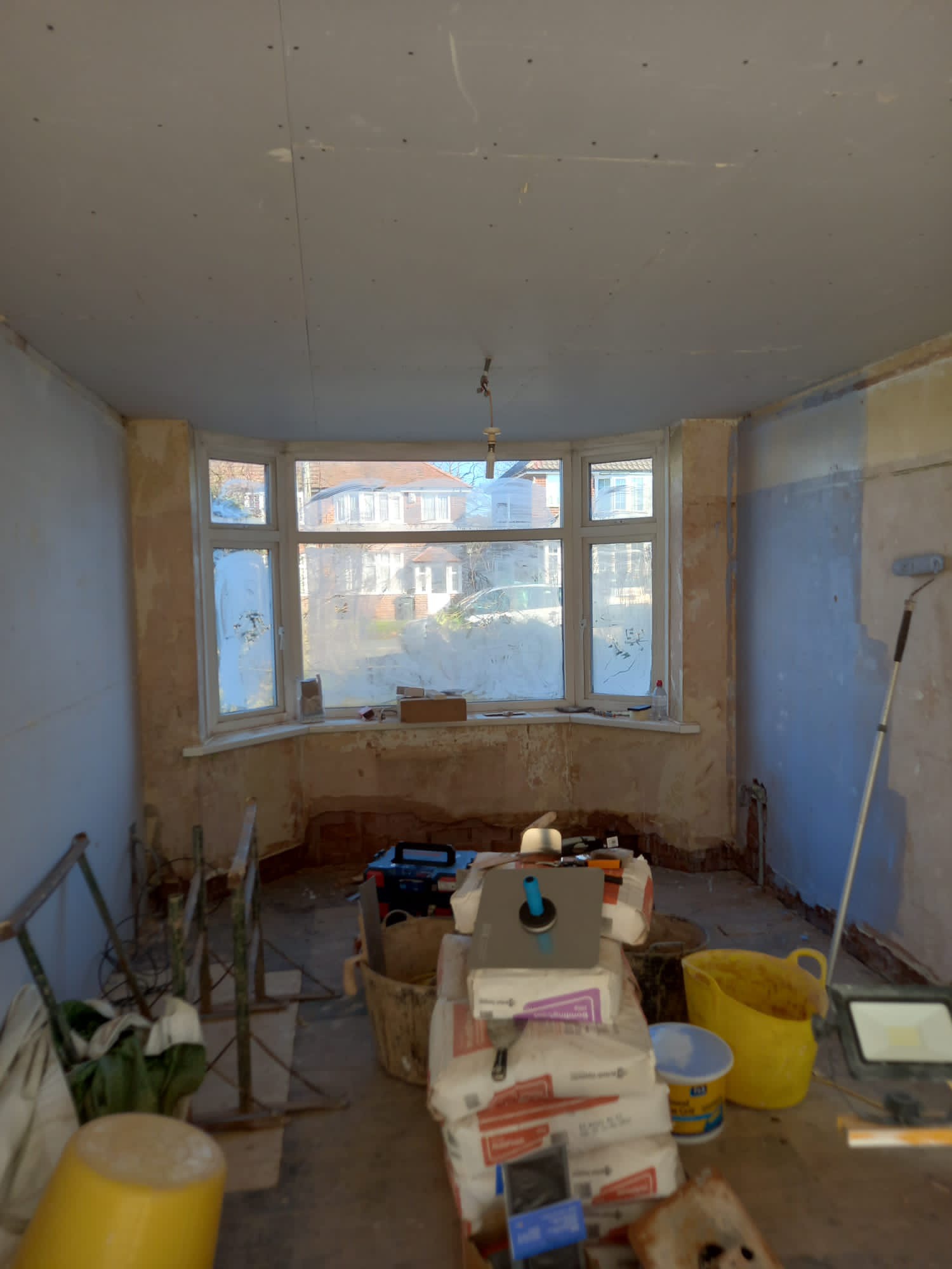 Pokój w trakcie przygotowania do remontu, z oknem wykuszowym, widoczne worki z zaprawą, wiadro, drabiny malarskie i częściowo pomalowana ściana na niebiesko.