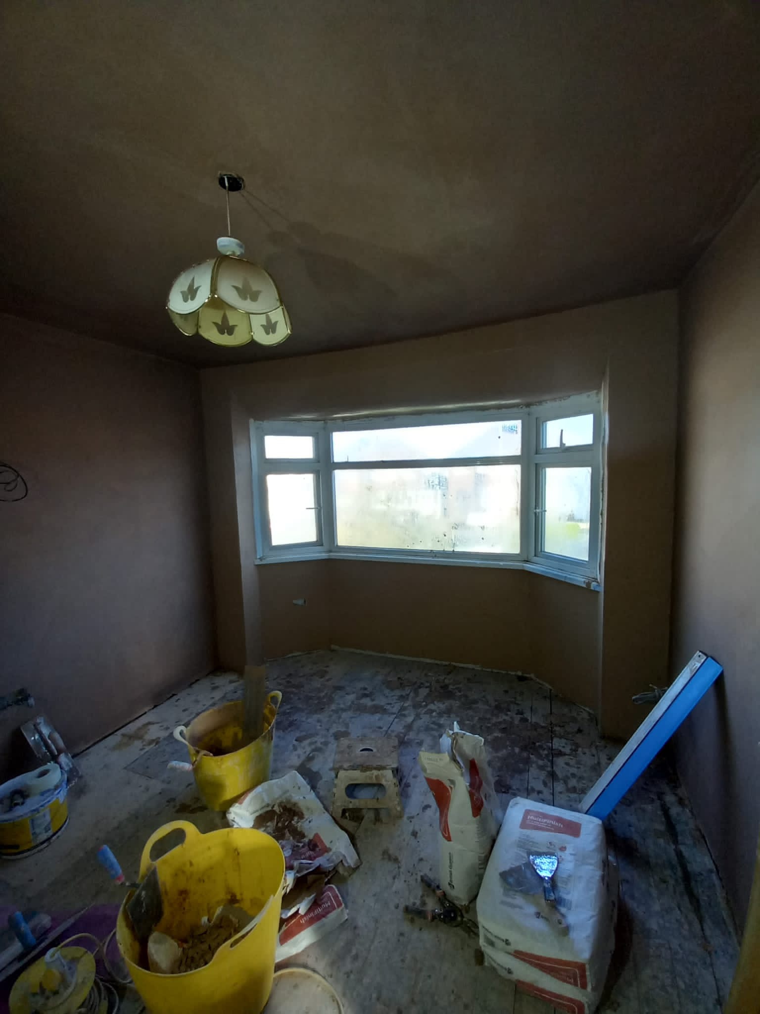 Pokój w trakcie prac wykończeniowych, ściany pokryte świeżym tynkiem, widoczne wiadra z zaprawą, worki z materiałem i narzędzia szpachlarskie na podłodze, okno wykuszowe z zaparowanymi szybami.