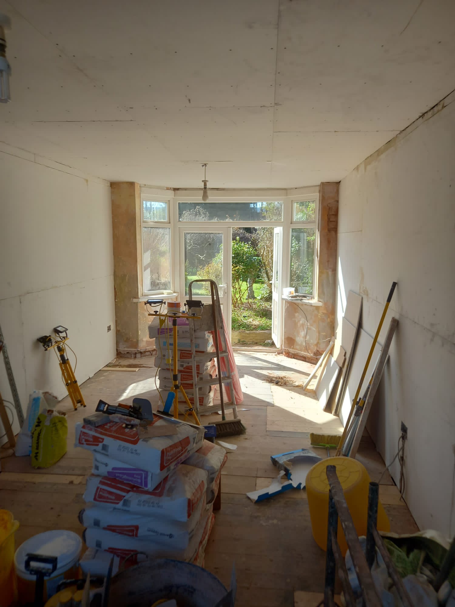 Pokój w trakcie przygotowania do remontu: ściany częściowo pokryte płytami gipsowymi, sterta worków z zaprawą, drabina oparta o okno balkonowe z widokiem na ogród, widoczne narzędzia i reflektory.