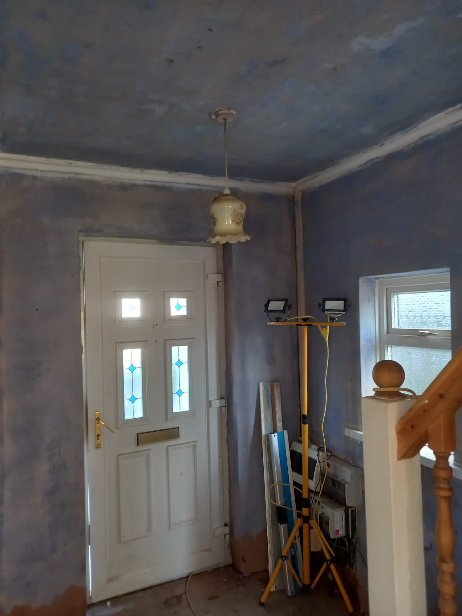 Wnętrze w trakcie przygotowania ścian do malowania: surowe ściany po szpachlowaniu, widoczny halogen na statywie, drzwi wejściowe z szybkami i schody z drewnianą balustradą.