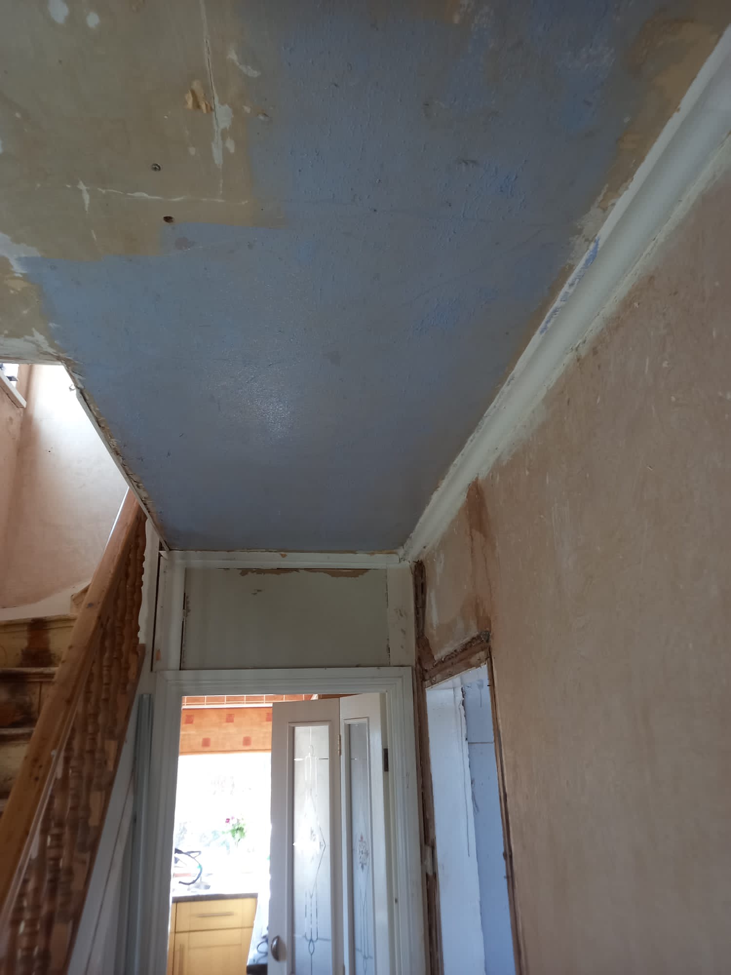 Klatka schodowa w trakcie przygotowania do malowania: widoczne ślady po starych tapetach, częściowo zagruntowany sufit na niebiesko, listwa przysufitowa, drewniane schody i futryna drzwi do kuchni...