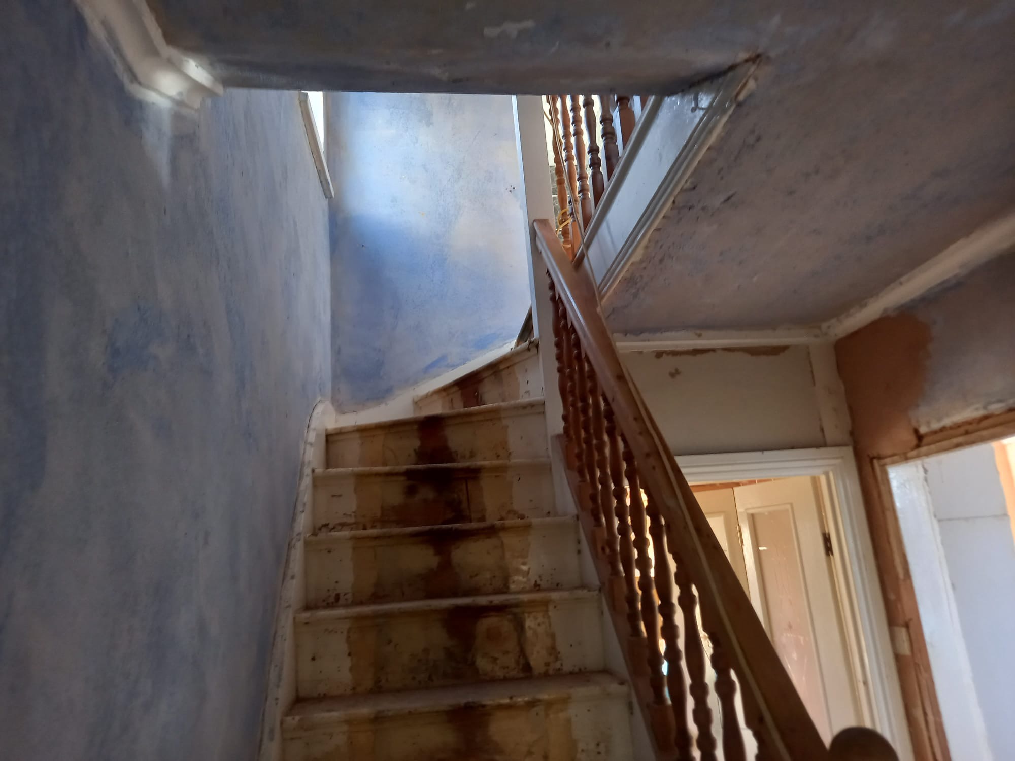Klatka schodowa w kamienicy w trakcie remontu: widoczne ślady po starych farbach na schodach i ścianach, sufit z odchodzącą farbą, drewniana balustrada, drzwi w tle.