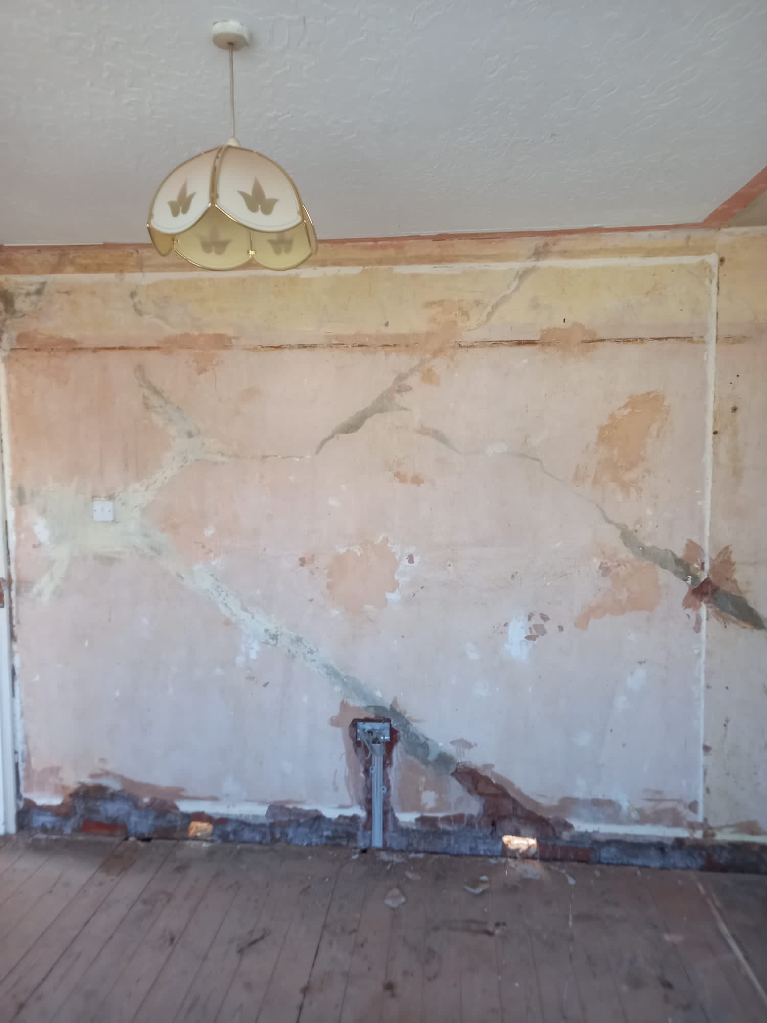Pokój w trakcie przygotowań do remontu: ściany z widocznymi pęknięciami i ubytkami, odsłonięte fragmenty muru przy podłodze, drewniana podłoga, lampa wisząca w stylu retro.