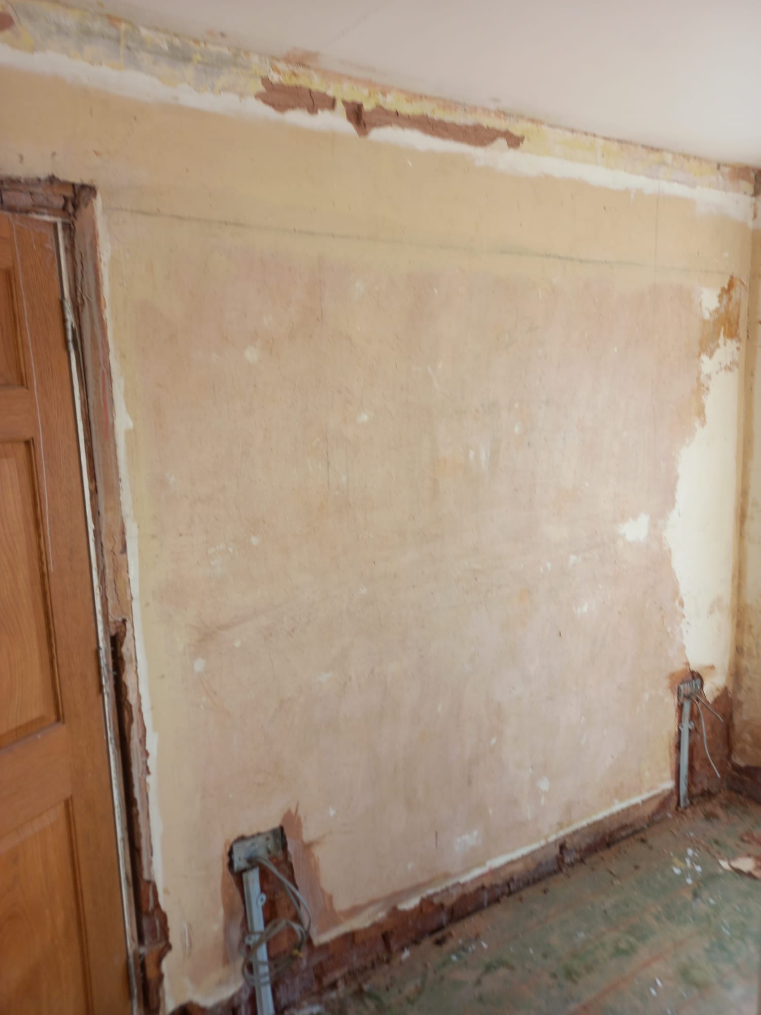 Przygotowana do szpachlowania ściana z widocznymi instalacjami elektrycznymi i uszkodzonymi fragmentami tynku przy listwach podłogowych i ościeżnicy drzwiowej.