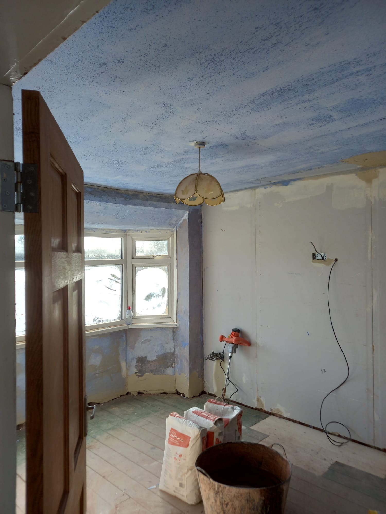 Wnętrze pokoju w trakcie przygotowania do malowania: sufit pokryty wstępną warstwą niebieskiej farby, ściany częściowo wyszpachlowane, widoczne worki z zaprawą, mieszadło elektryczne i otwarte drzwi.