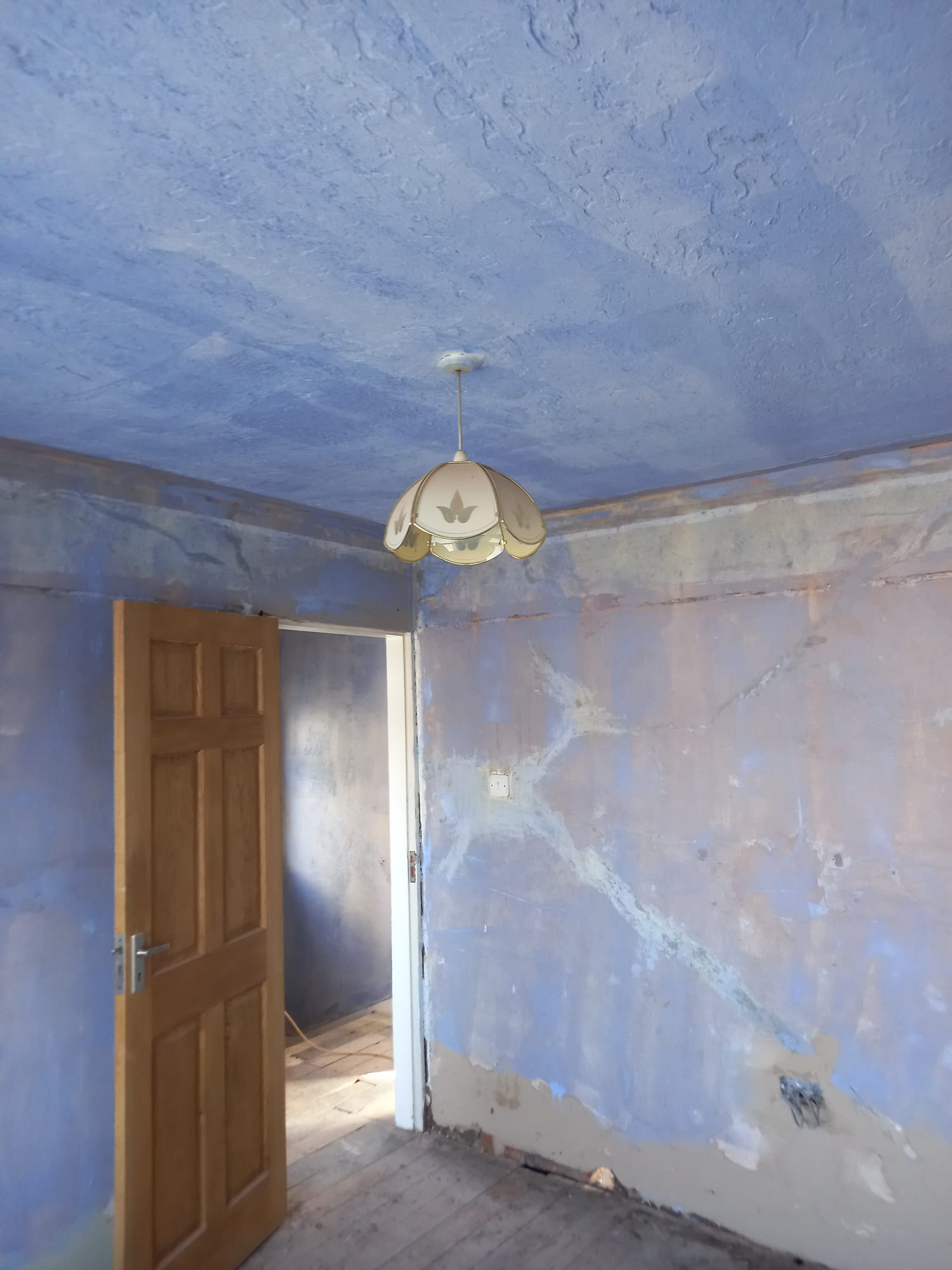 Pokój w trakcie przygotowań do malowania: ściany i sufit pokryte niebieską farbą z widocznymi śladami szpachlowania, wisząca lampa z kloszem, otwarte drewniane drzwi i fragment podłogi.
