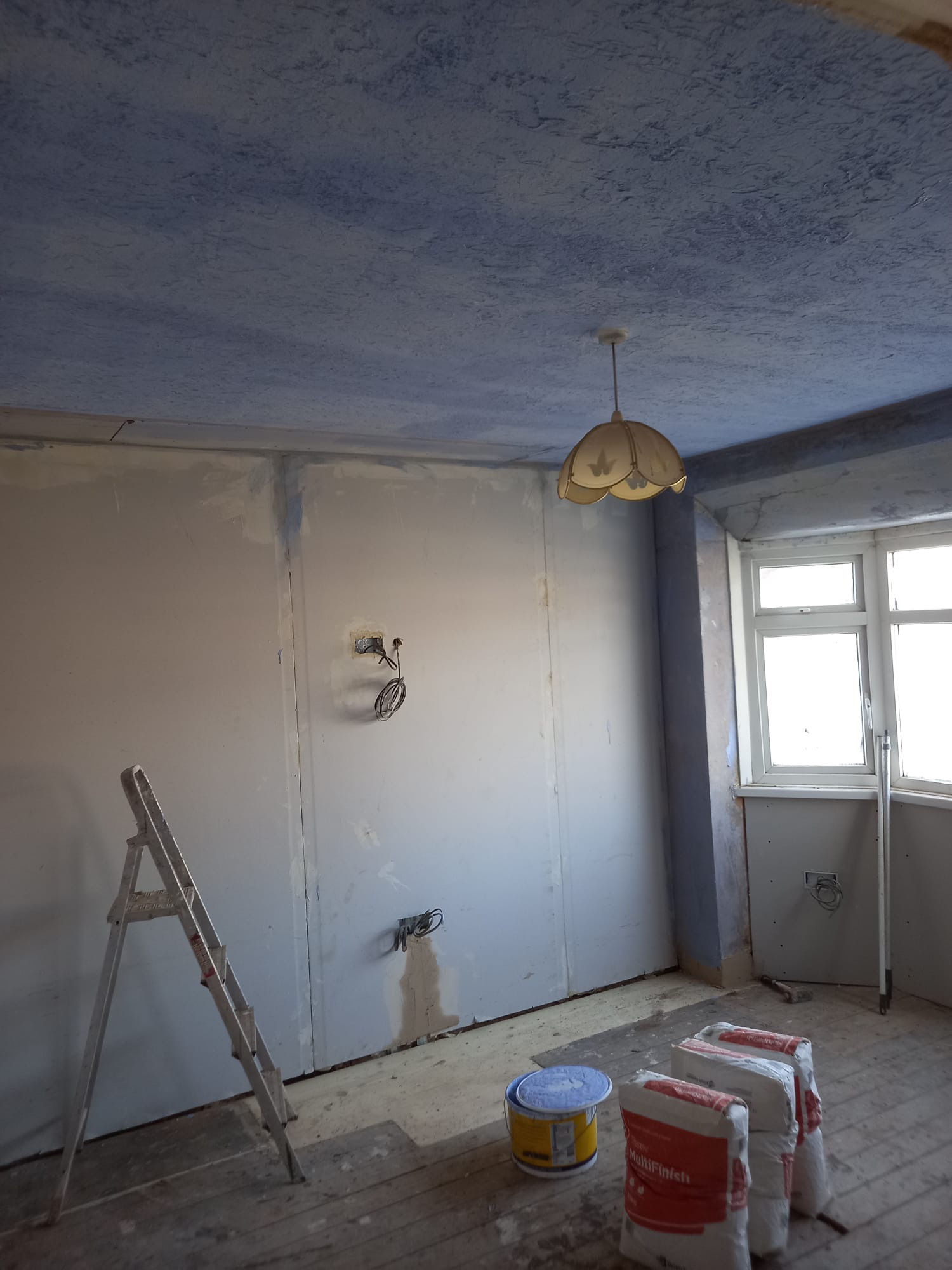 Pokój w trakcie prac wykończeniowych: pomalowany na niebiesko sufit z fakturą, ściany przygotowane do dalszej obróbki, drabina, wiadro z farbą i worki z gładzią na podłodze z desek.
