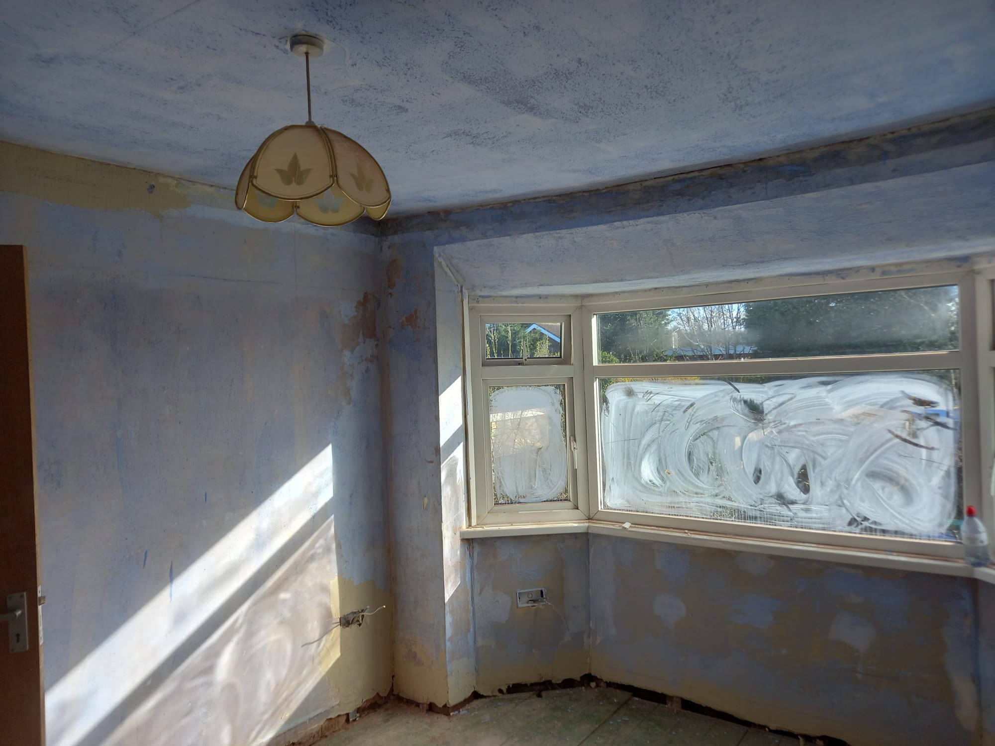 Pokój w trakcie remontu: ściany częściowo wyszpachlowane, okno z zabrudzonymi szybami, odsłonięte przewody elektryczne, stara lampa wisząca.