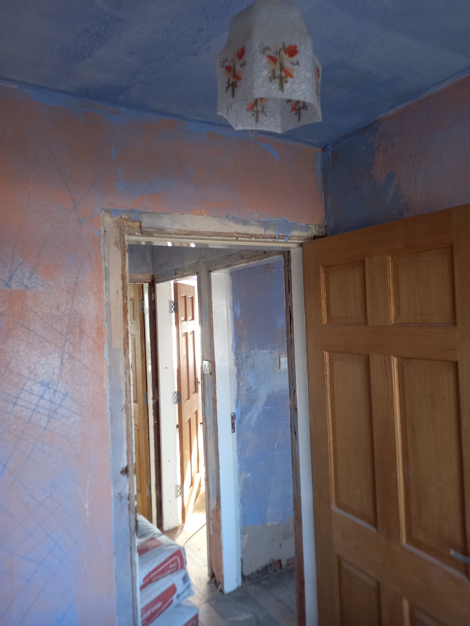 Wnętrze w trakcie przygotowania do remontu: ściany z widocznymi śladami szpachlowania i zarysowanymi liniami, worki z materiałem budowlanym w rogu, drewniane drzwi w ościeżnicy i lampa sufitowa...