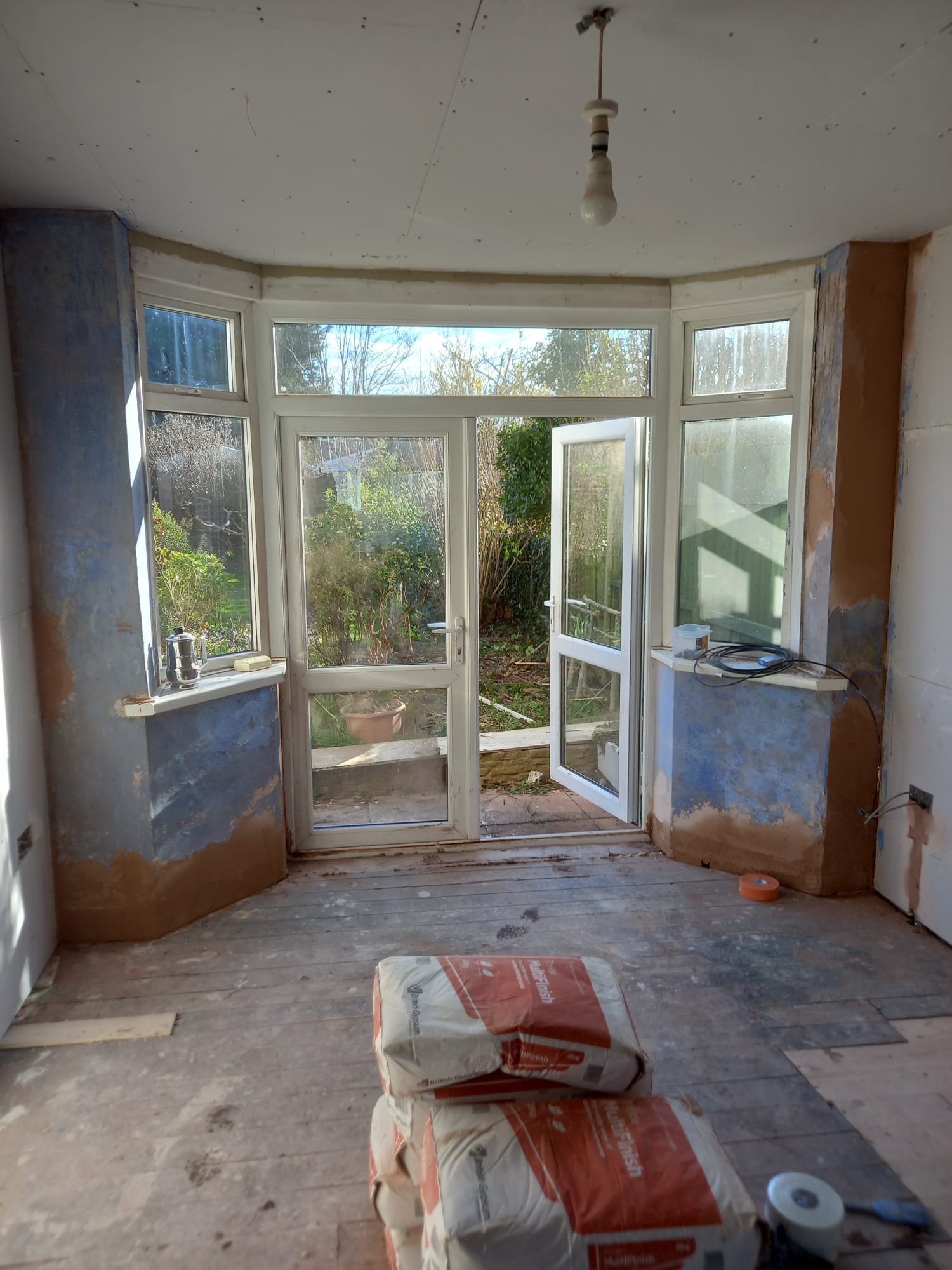 Pokój w trakcie remontu z widocznymi workami gładzi, surowymi ścianami z częściowo nałożoną warstwą, drewnianą podłogą i otwartymi drzwiami balkonowymi wychodzącymi na ogród.