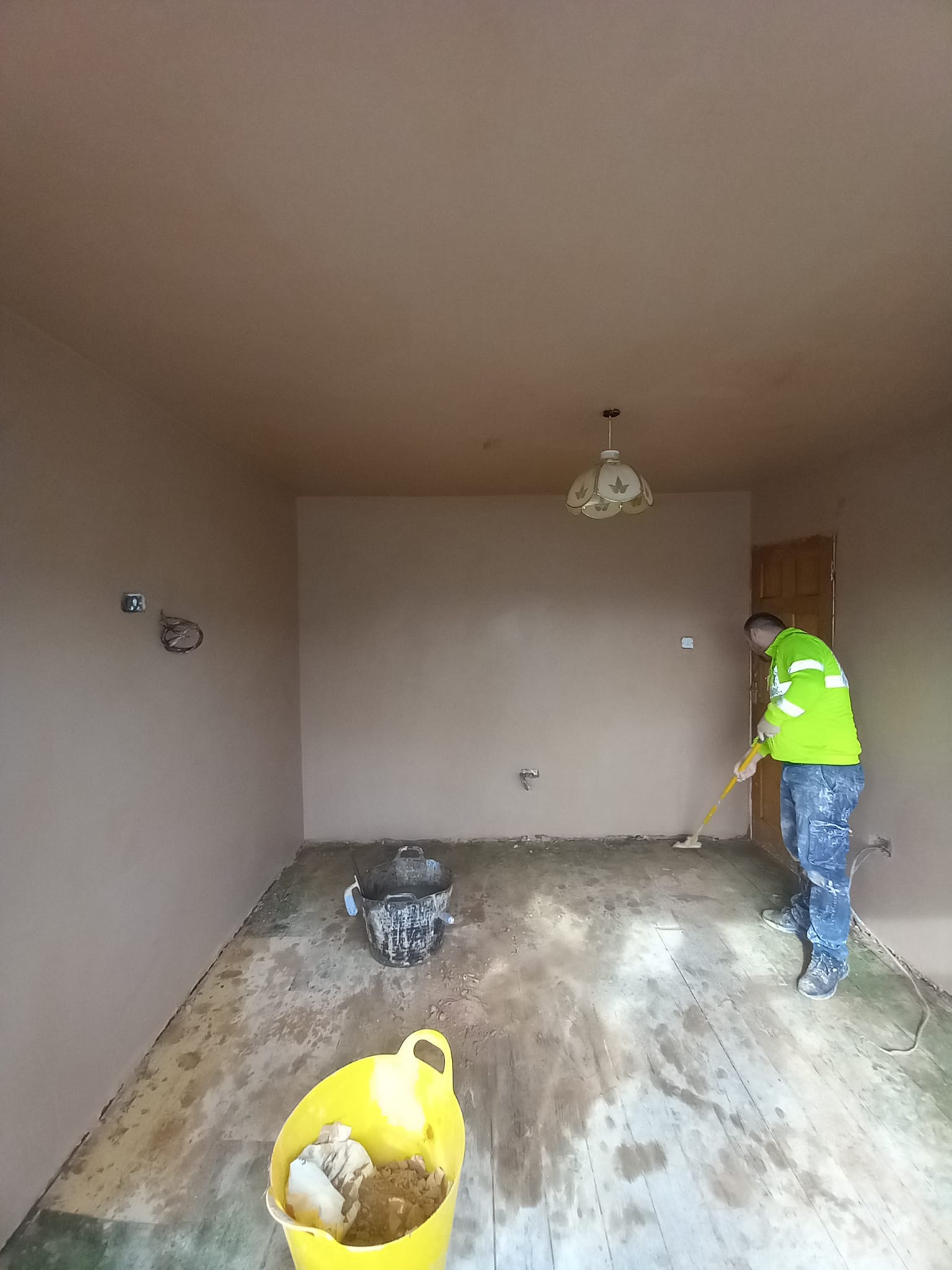 Pracownik w odblaskowej kurtce wygładza szpachlą podłogę w pokoju po nałożeniu gładzi na ściany, widoczne dwa wiadra z materiałami.