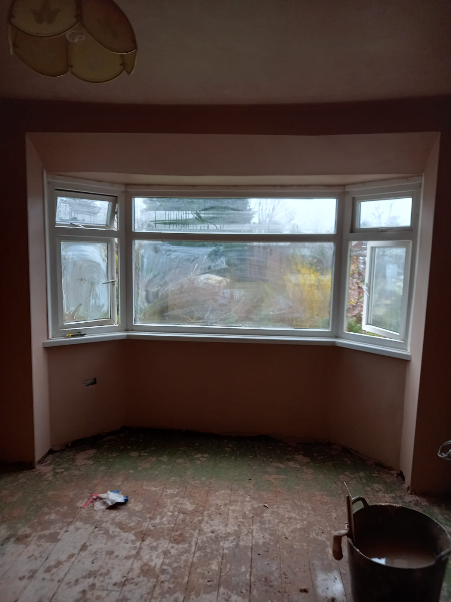 Pokój w trakcie prac wykończeniowych: ściany po szpachlowaniu, brudne okna z widokiem na ogród, drewniana podłoga w stanie surowym, wiadro z zaprawą w rogu.