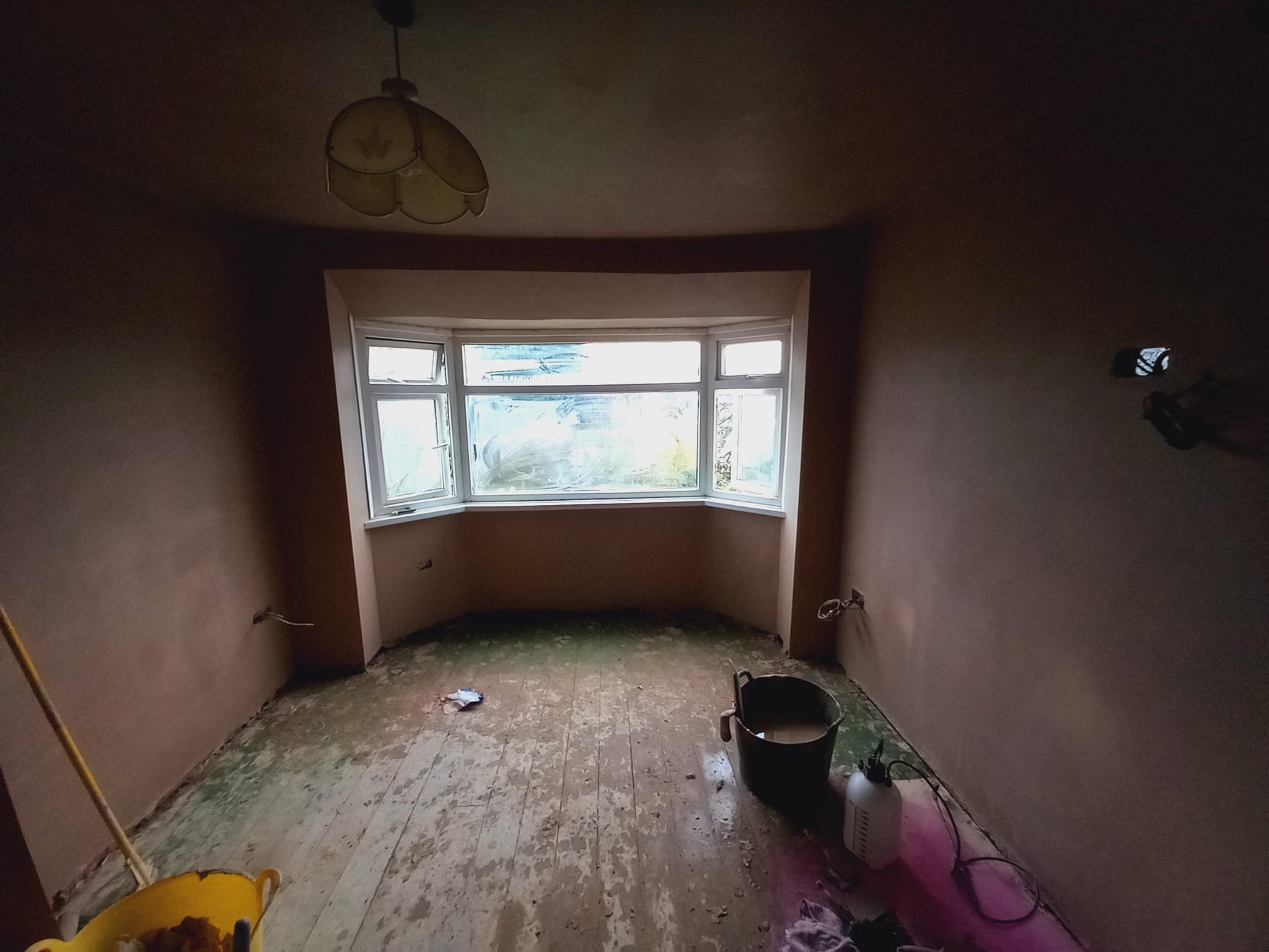 Pokój po szpachlowaniu ścian, z widocznymi śladami gładzi na podłodze i narzędziami pracy, okno wykuszowe w tle.