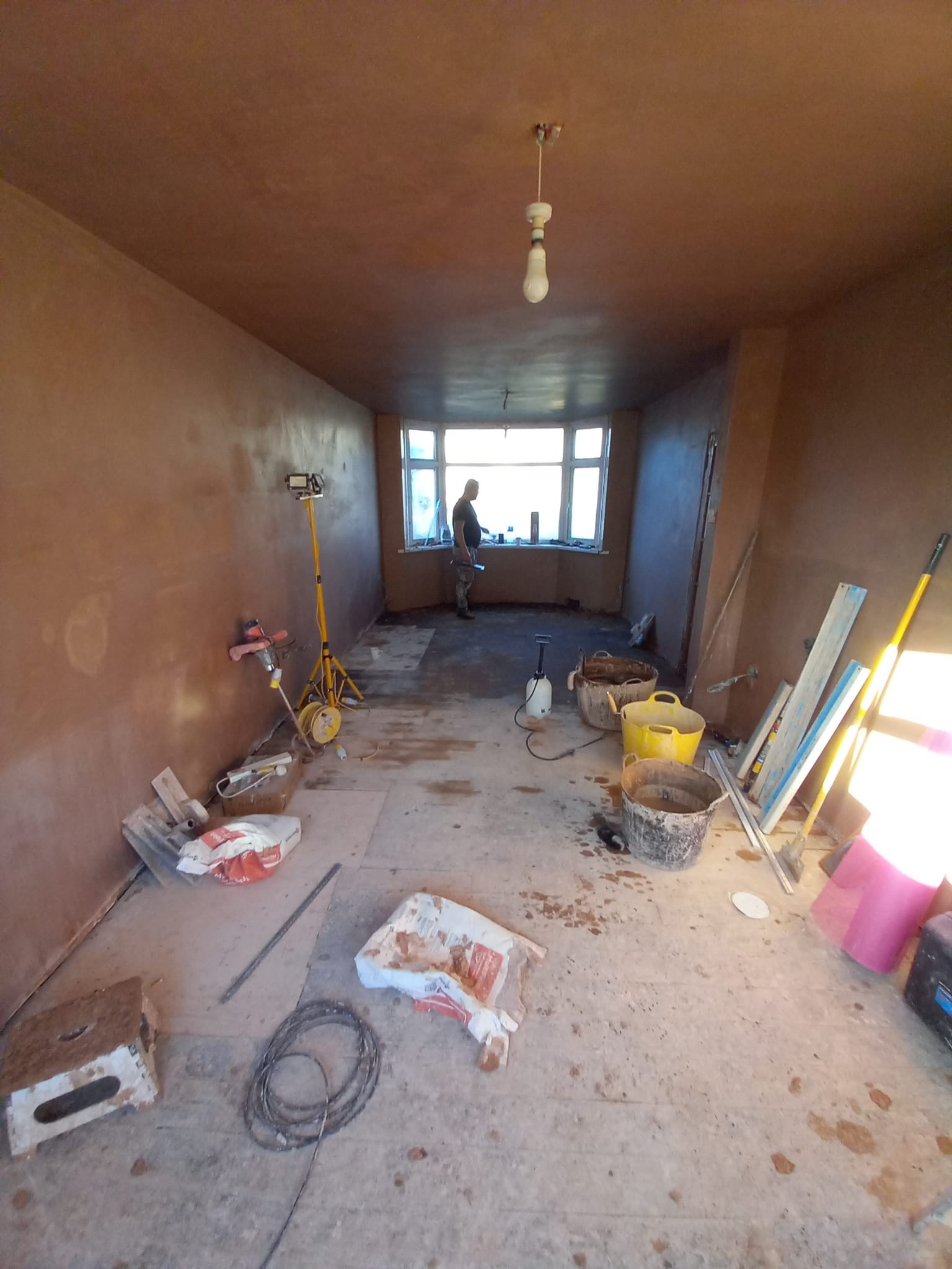 Pokój w trakcie szpachlowania ścian, z widocznymi narzędziami, wiadrami i workami gipsu, oświetlony halogenem na trójnogu, z sylwetką osoby w oknie.