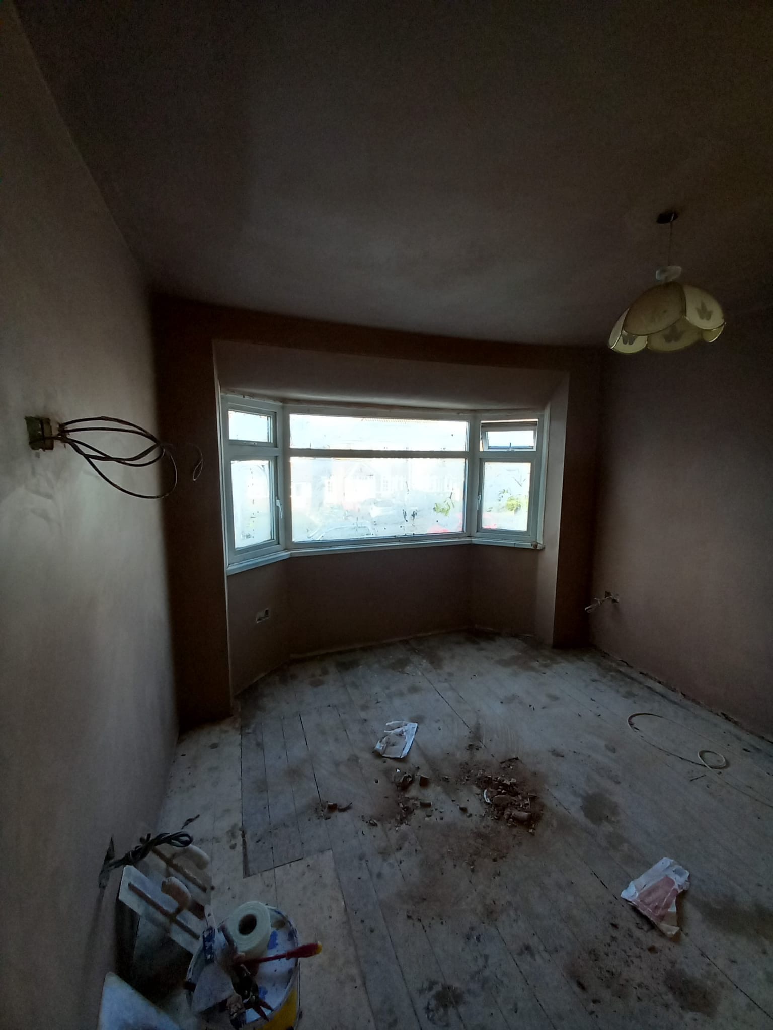 Pokój w trakcie przygotowania do prac wykończeniowych: świeżo otynkowane ściany, odsłonięte przewody elektryczne, drewniana podłoga z resztkami gruzu i narzędzia malarskie w rogu.
