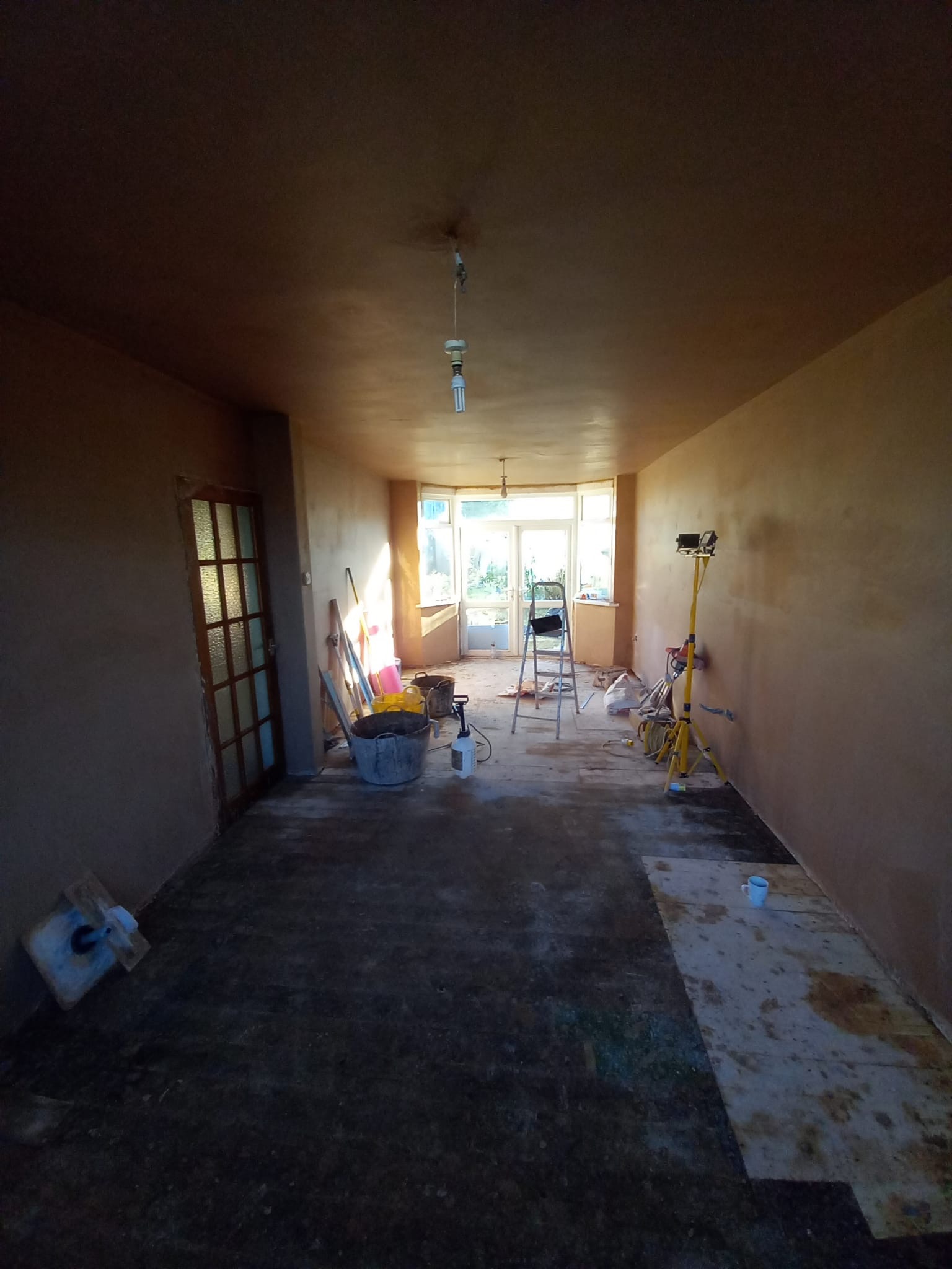 Pokój w trakcie prac wykończeniowych: świeżo nałożona gładź na ścianach i suficie, widoczne narzędzia, drabina, wiadro z zaprawą i lampa budowlana, kubek na podłodze, światło słoneczne wpadające...