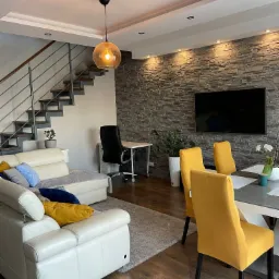 Jasne wnętrze salonu z białym sk&oacute;rzanym narożnikiem, szarym dywanem, schodami z metalową balustradą, ścianą z kamienia dekoracyjnego, telewizorem, ż&oacute;łtymi krzesłami i stołem, oświetlone lampą...